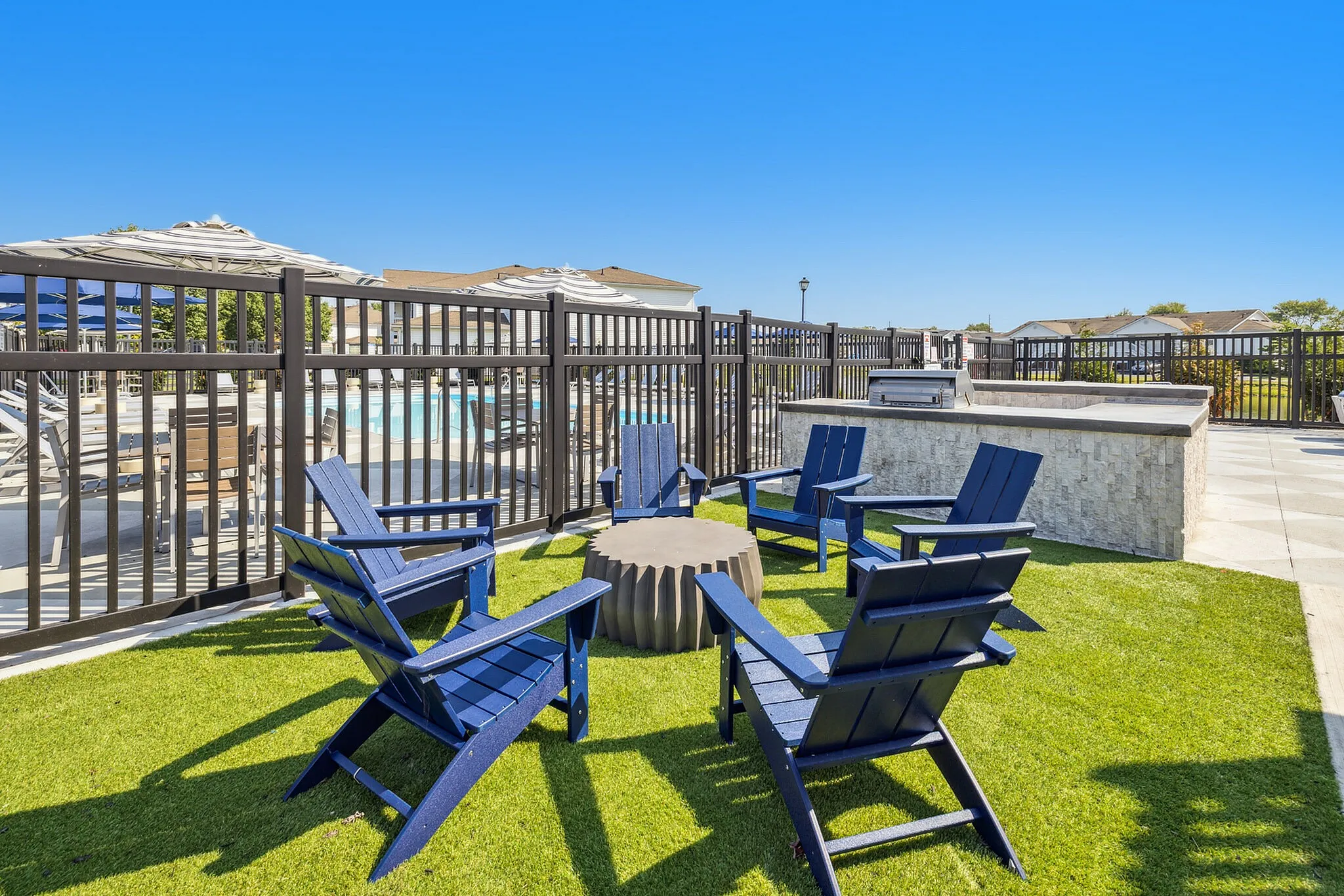 Inviting outdoor patio with stylish blue chairs around a fire pit, perfect for relaxation, adjacent to a modern BBQ station and overlooking a sparkling pool.