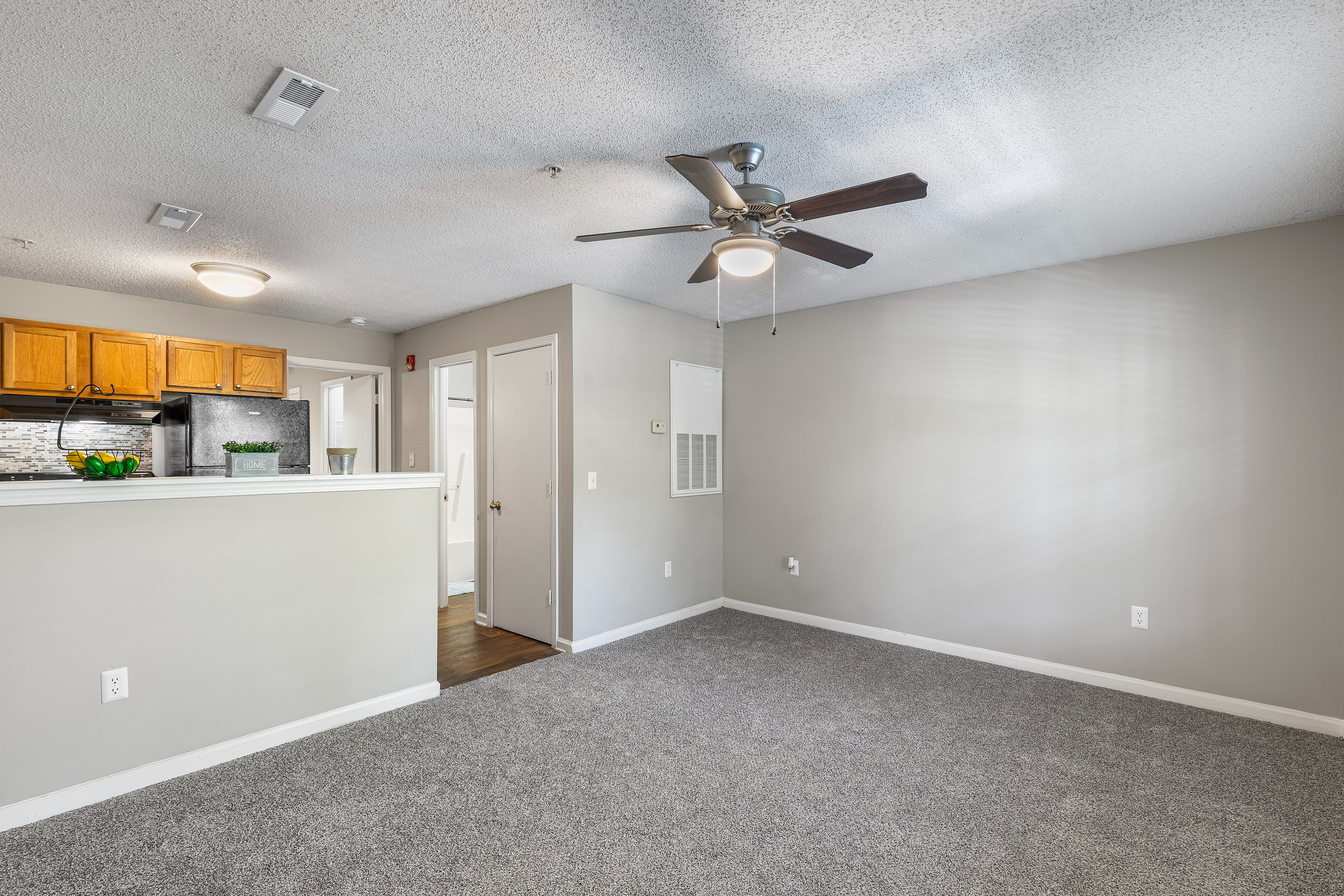 Inviting living space with textured carpet, modern ceiling fan, and an open kitchen featuring warm wooden cabinets and sleek countertops.