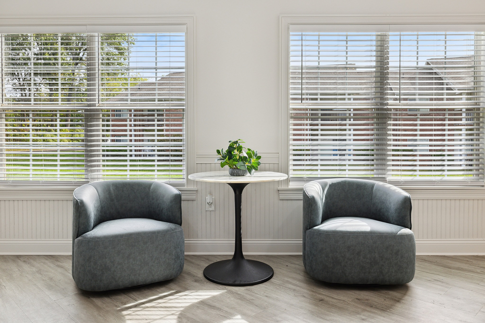 Elegant and bright seating area featuring two modern gray chairs, a sleek round table, and large windows with scenic outdoor views.