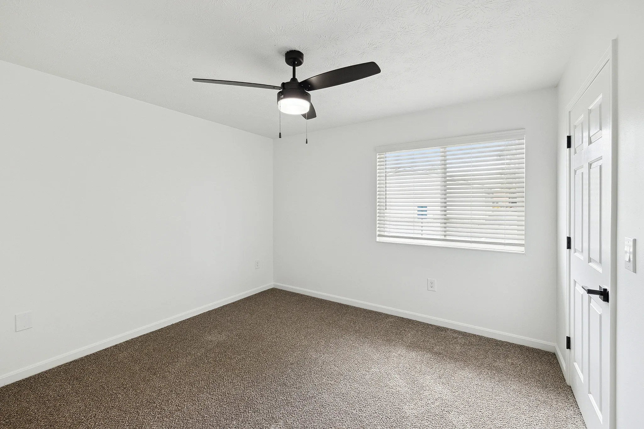 Bright and inviting room with modern ceiling fan, large window with blinds, and plush carpet flooring.