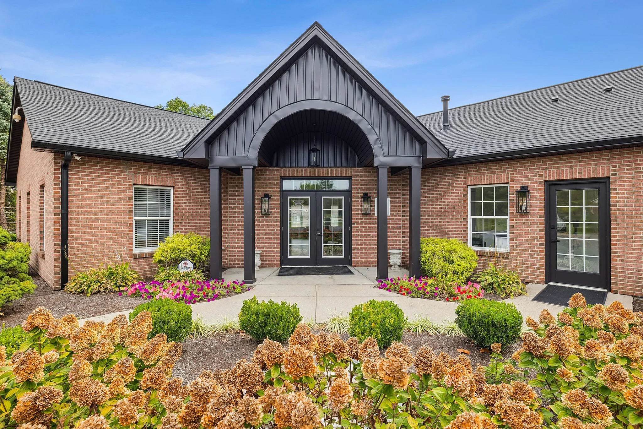 Charming brick building entrance with elegant black accents, surrounded by vibrant landscaping and colorful flowers, under a clear blue sky.