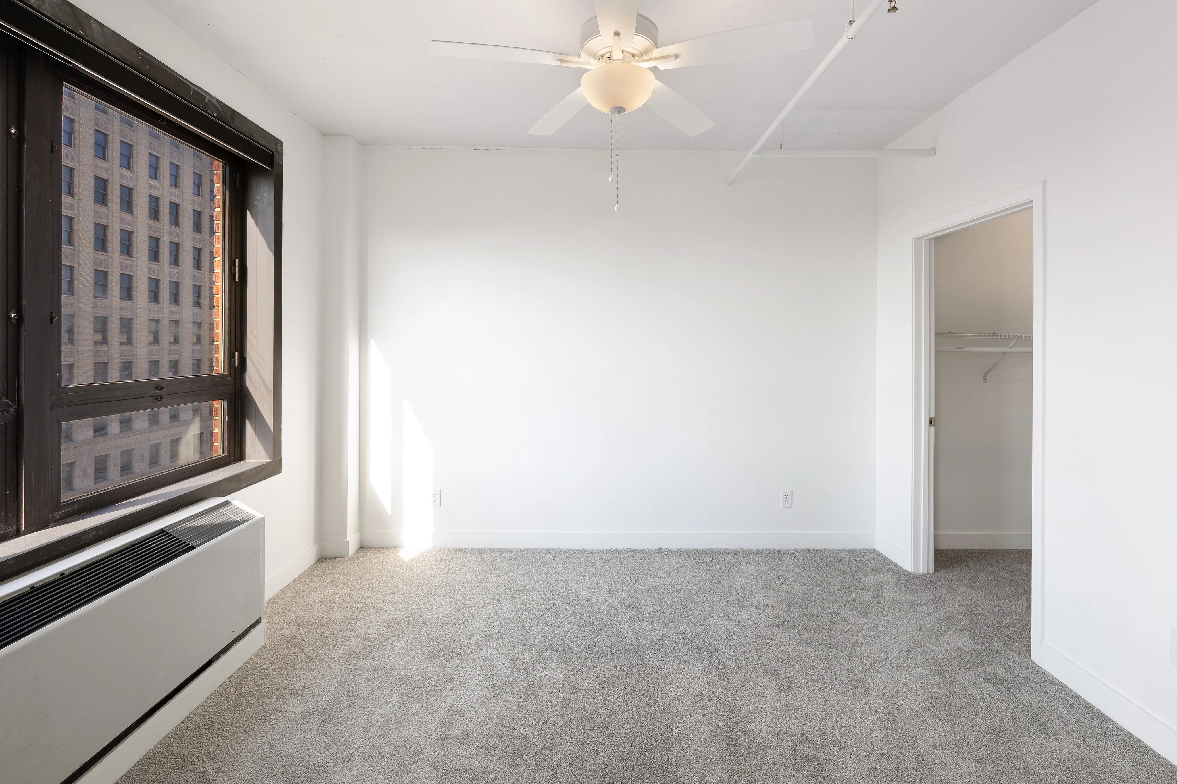 Bright, carpeted bedroom with large window and downtown city view, featuring a ceiling fan and a spacious walk-in closet.
