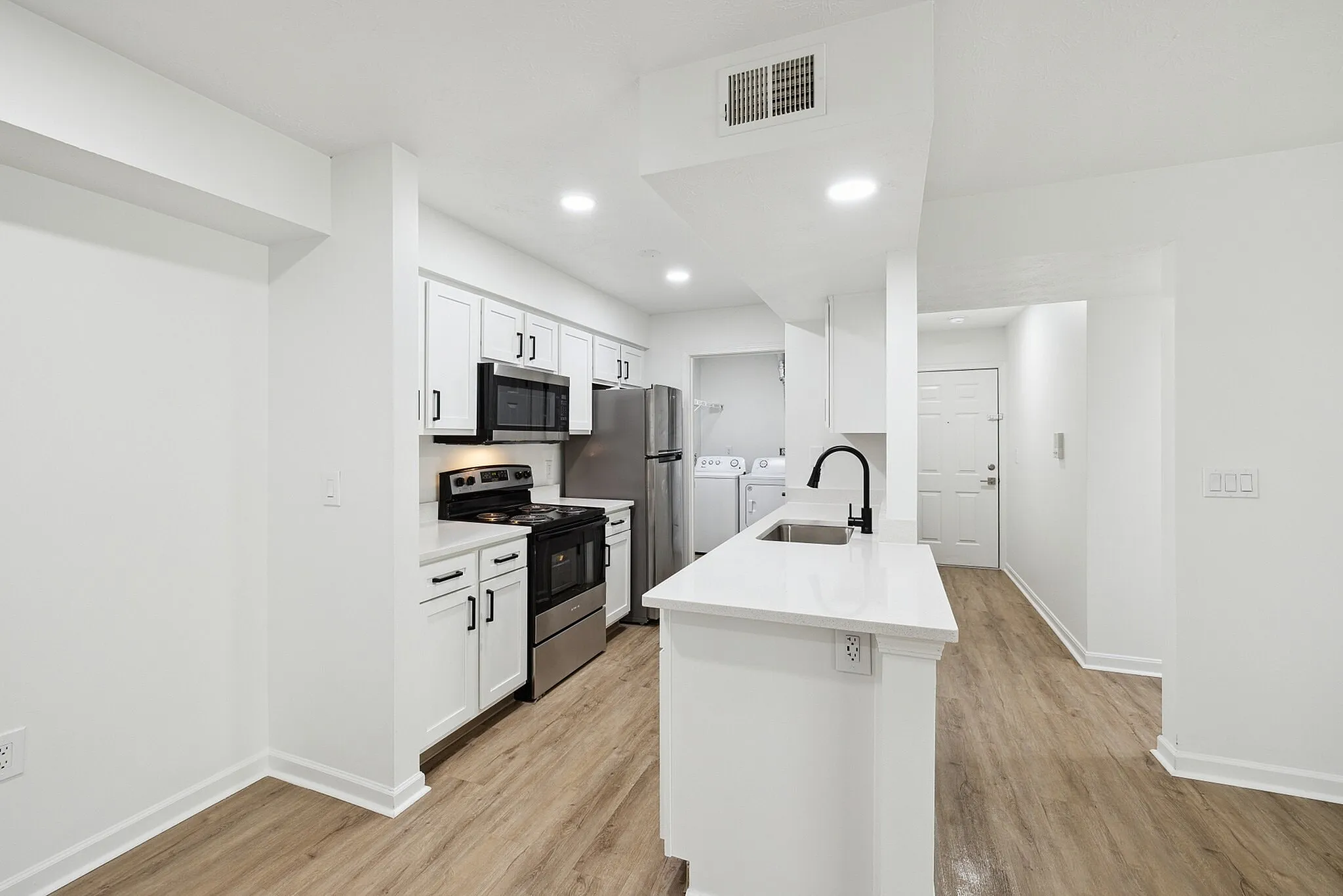 Modern kitchen boasting sleek white cabinetry, stainless steel appliances, and elegant wood flooring, leading to a convenient laundry area.
