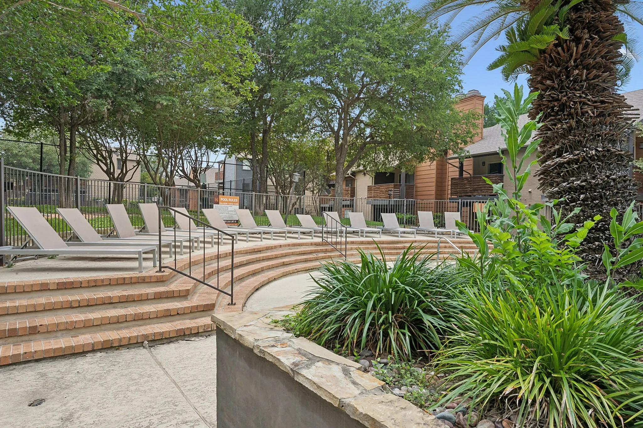 Inviting outdoor lounge area with sleek sun loungers, surrounded by lush greenery and tree-lined pathways.