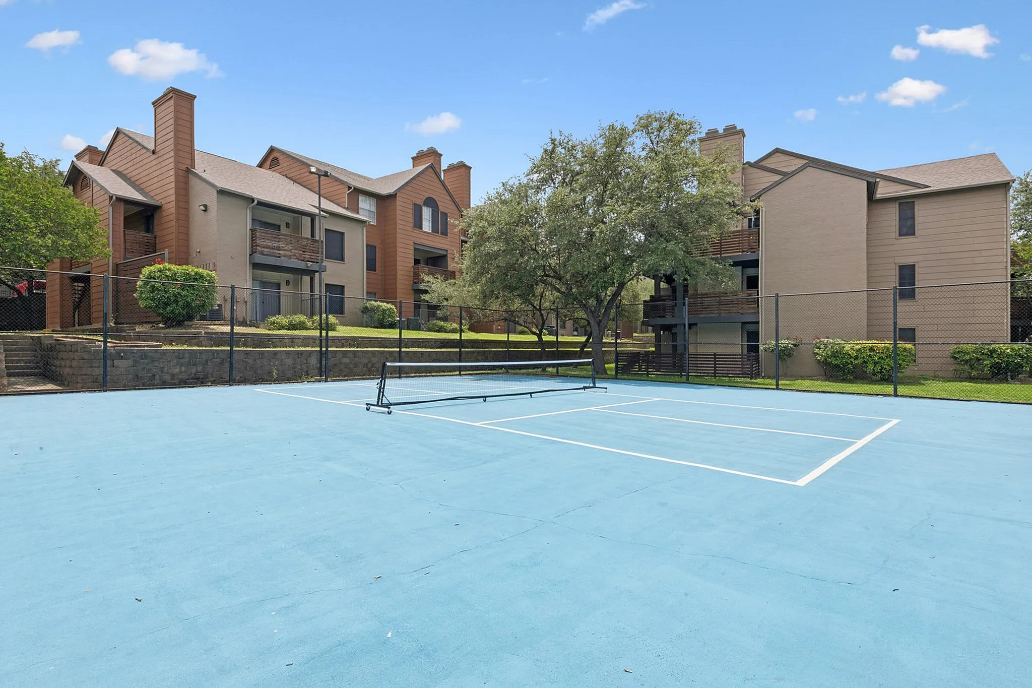 Sunny residential complex featuring charming brick buildings surrounded by lush greenery with a private blue tennis court.