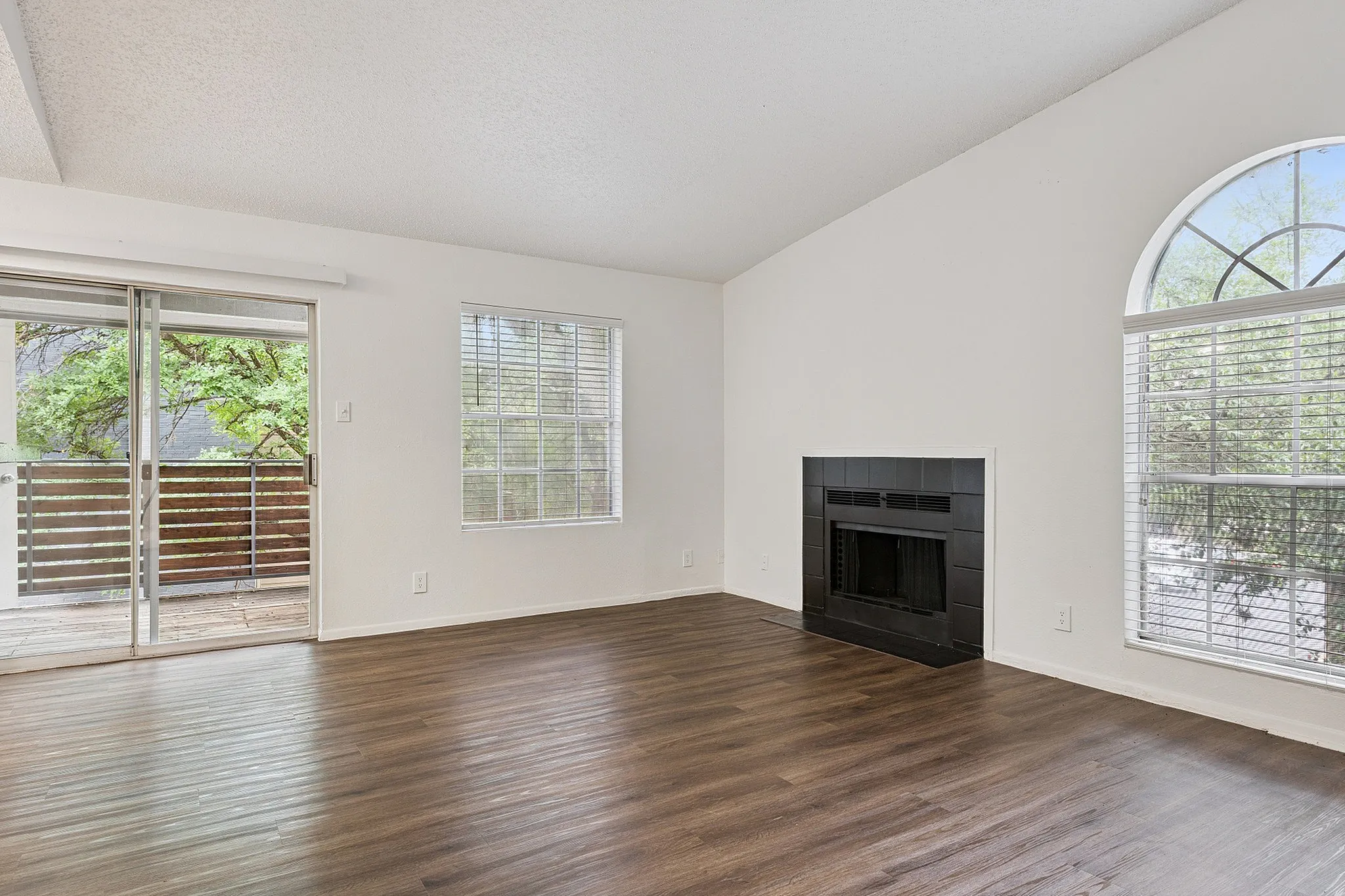 Bright living room with elegant hardwood floors, large windows for natural light, and a cozy fireplace, leading to a charming balcony with a view of lush greenery.