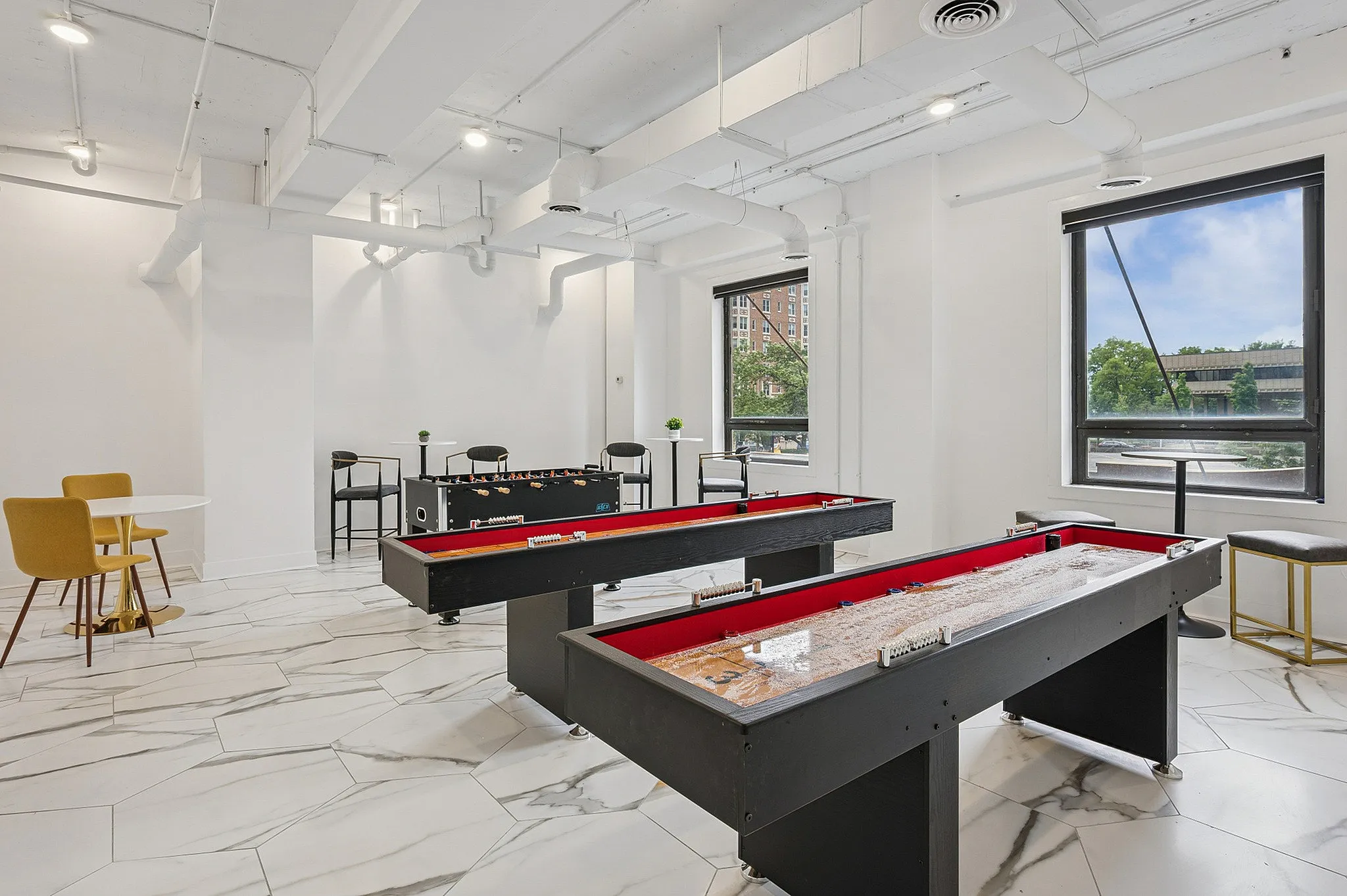 Bright and modern game room featuring shuffleboard and foosball tables, sleek white interiors, and stylish seating with large windows providing natural light.