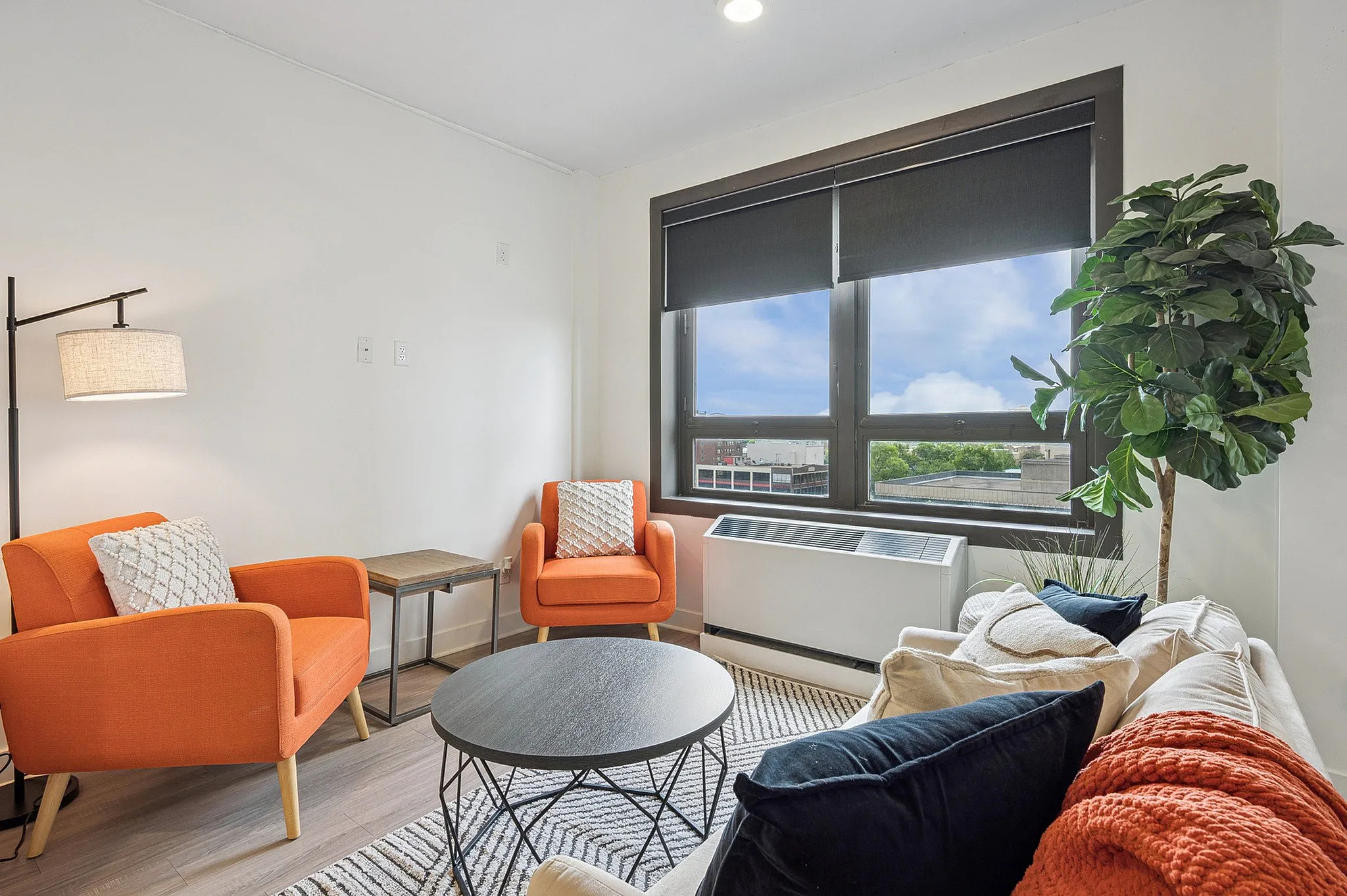 Bright and inviting living room featuring stylish orange armchairs, a cozy sofa with plush cushions, and a large window with city views, bathed in natural light.