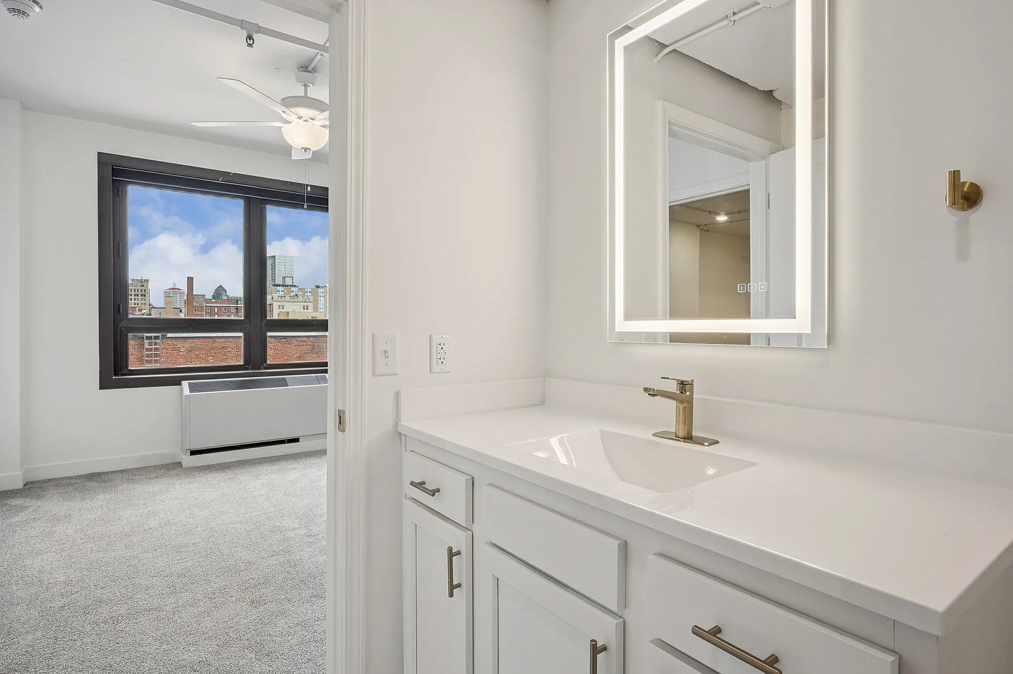 Modern apartment bathroom with elegant vanity and illuminated mirror, overlooking a captivating cityscape through large windows.