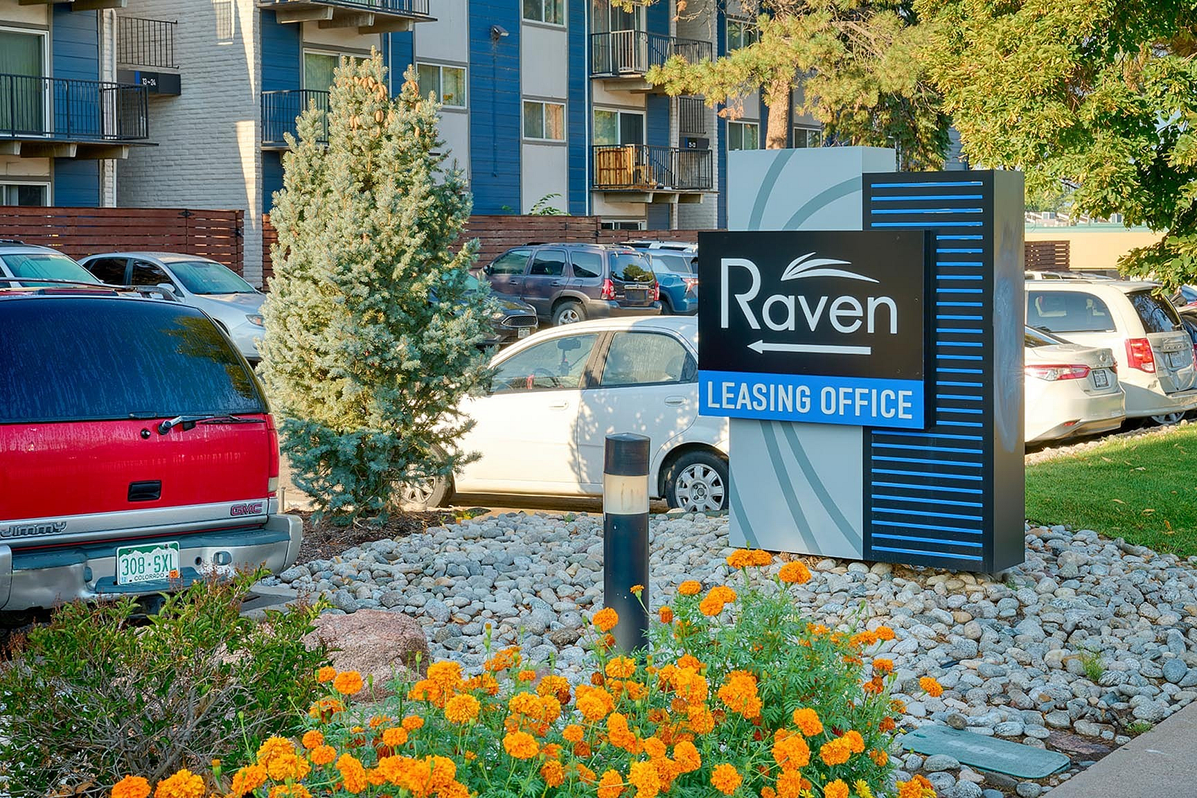 Inviting leasing office sign at Raven apartment complex, complemented by vibrant flowers and greenery. Modern building and ample parking visible in the background.