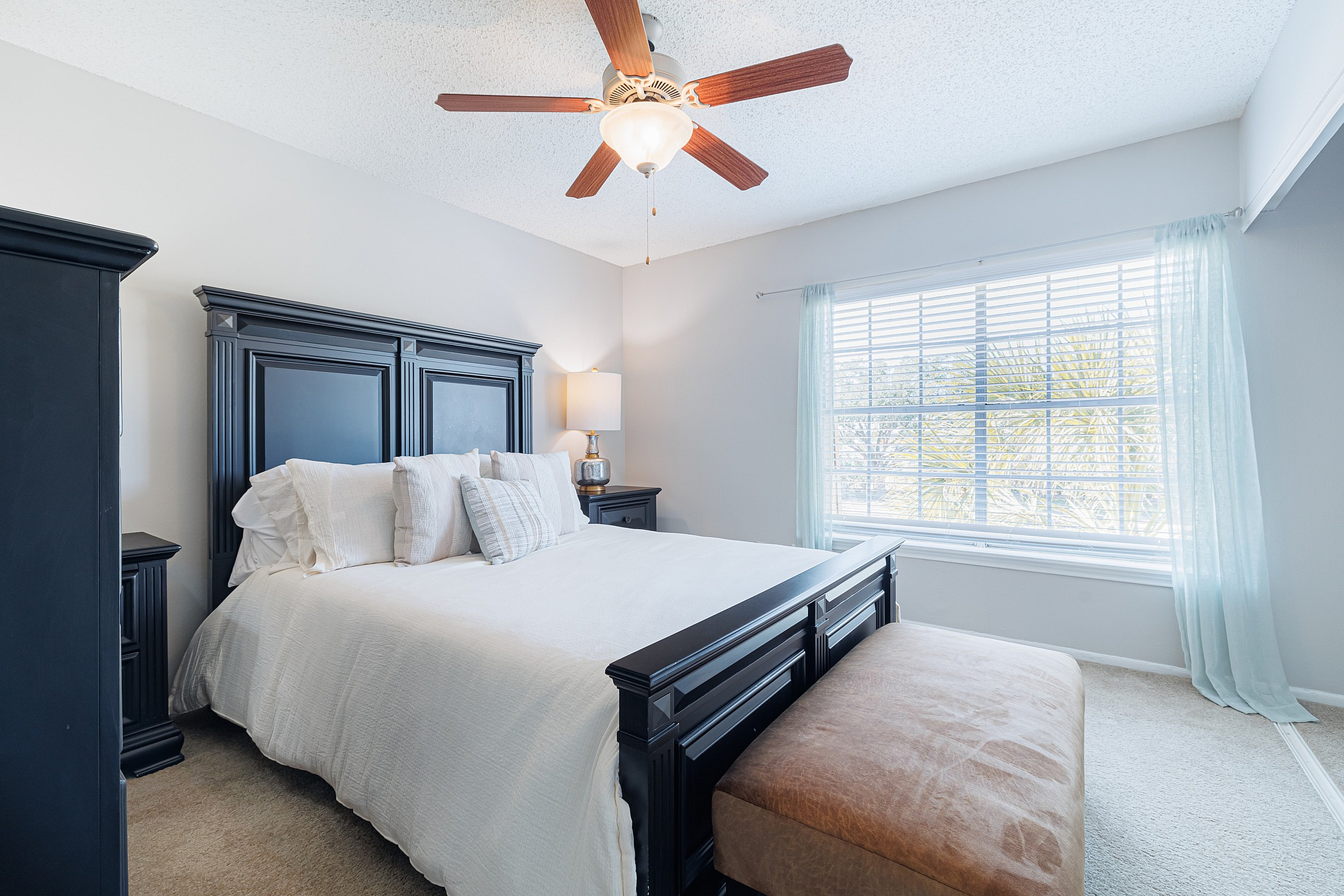 Bright and inviting bedroom with elegant dark wood furniture, luxurious bedding, ceiling fan, and a sunlit window creating a serene atmosphere.