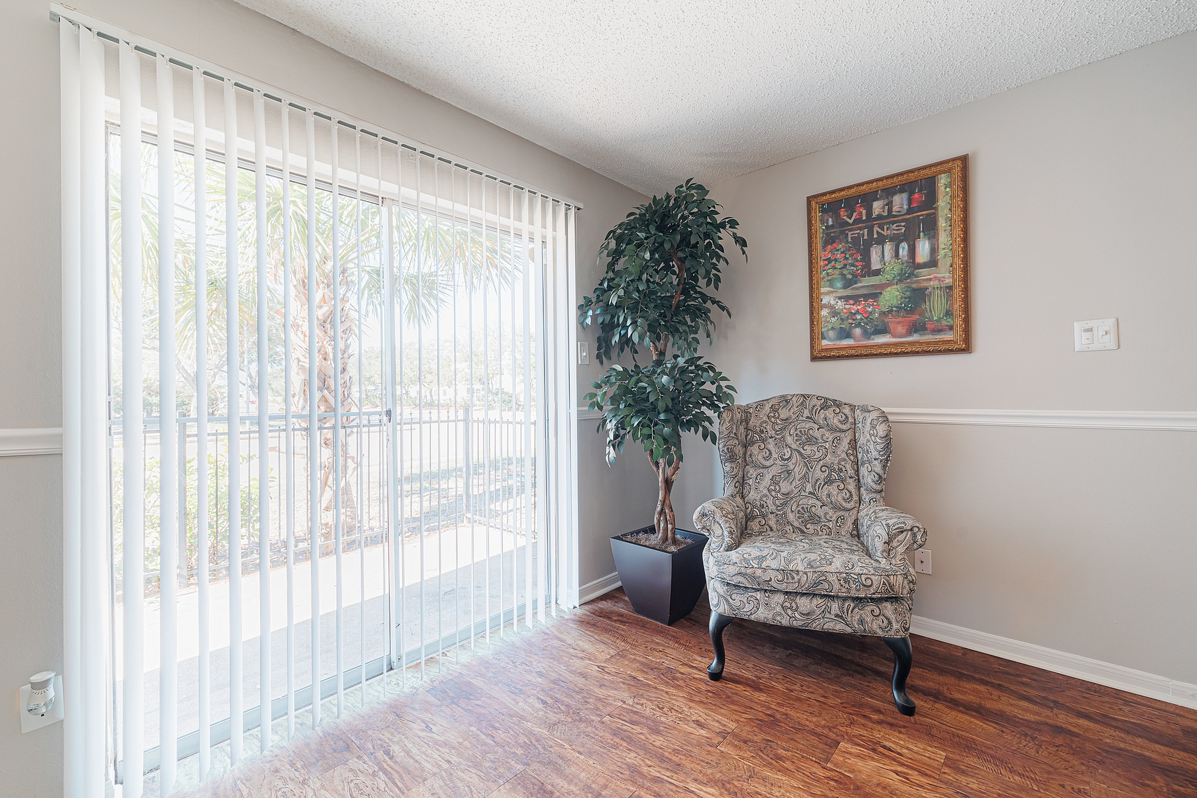 Bright and inviting corner with a stylish armchair, decorative plant, and large window featuring vertical blinds, overlooking a lush outdoor view.