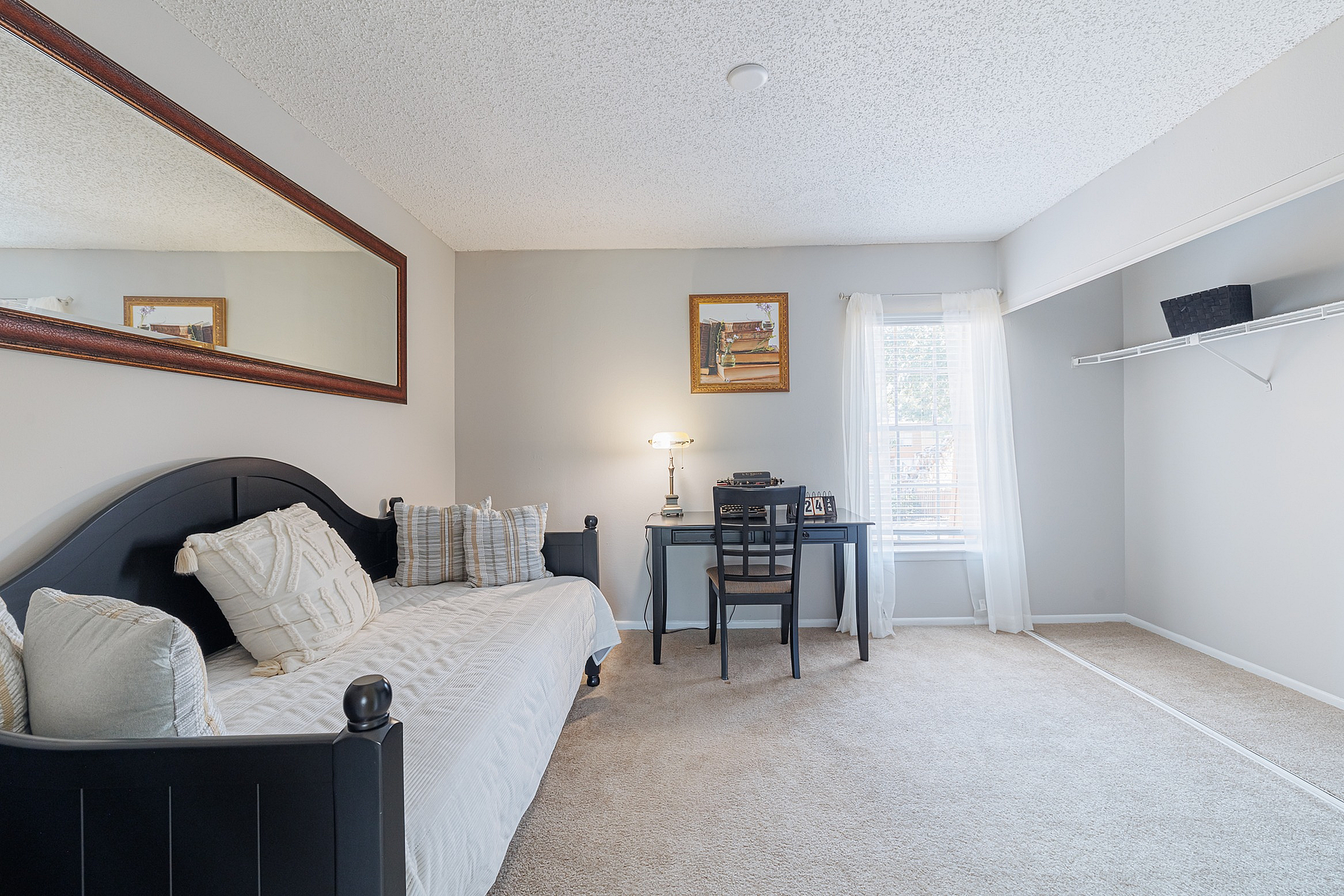 Elegant bedroom featuring a cozy daybed with plush pillows, a modern desk with a lamp, and a large mirror, bathed in natural light from a sunlit window.