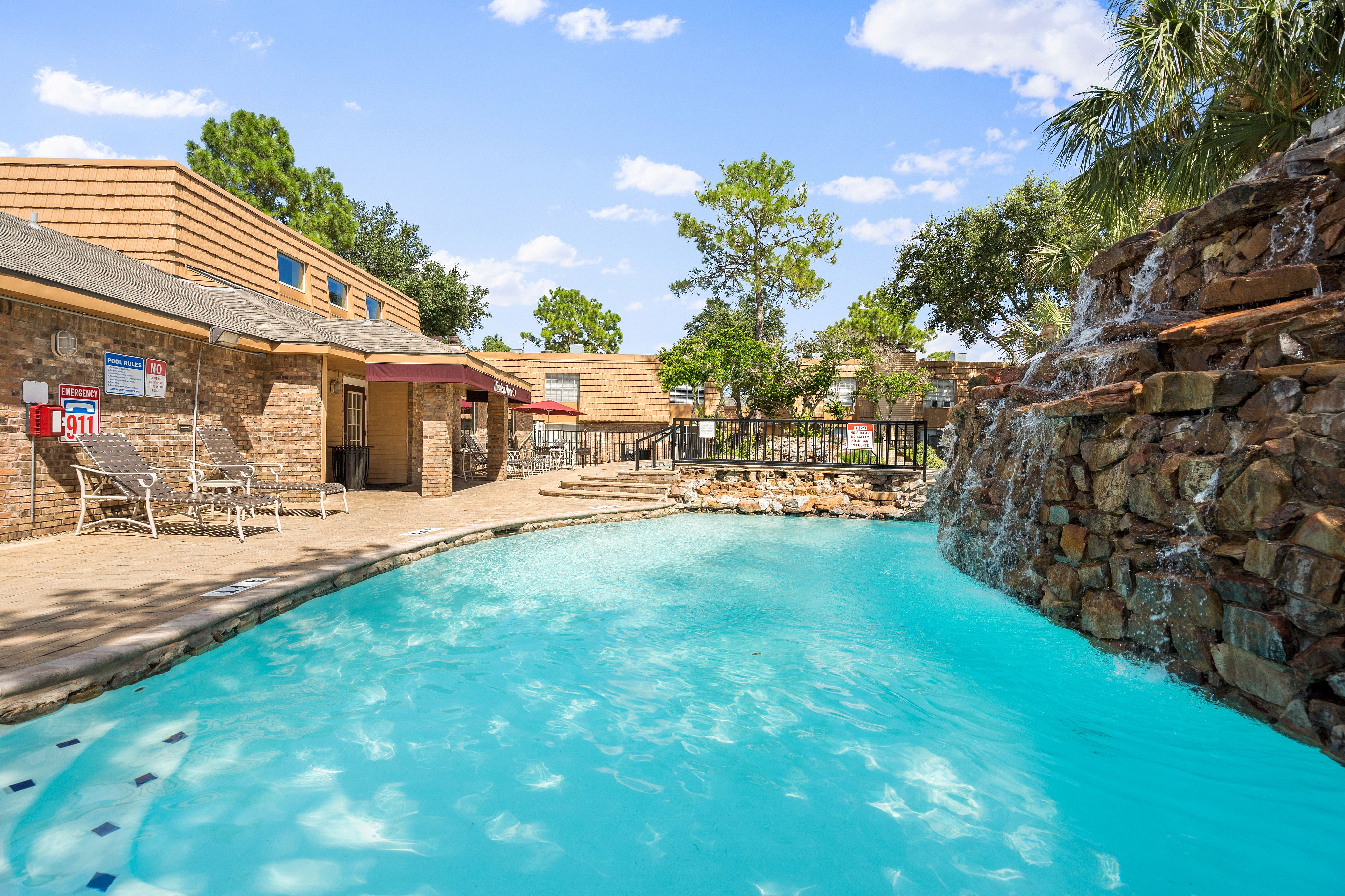 Inviting apartment complex pool area with a stunning waterfall feature and comfortable lounging chairs, surrounded by lush greenery and classic brick architecture.