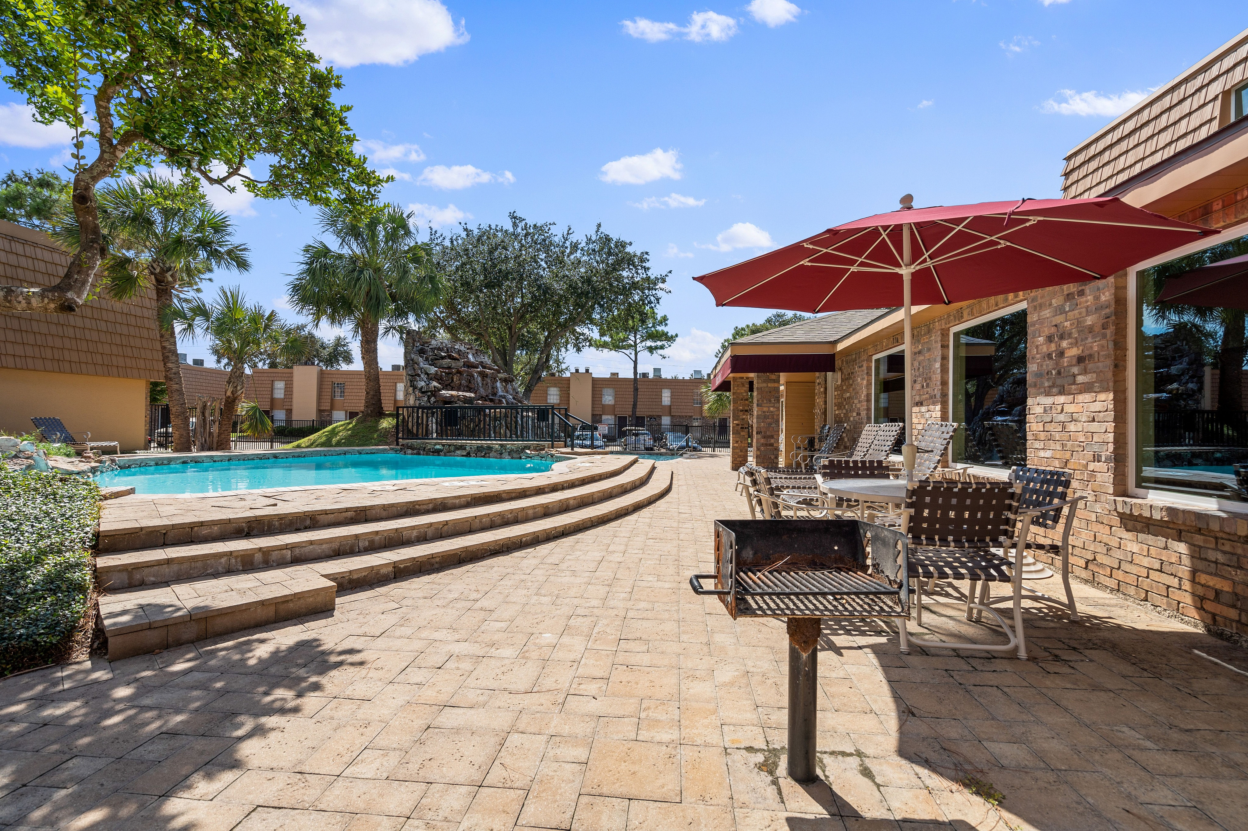 Inviting outdoor area featuring a sparkling pool, shaded patio with red umbrellas, and lush palm trees, perfect for relaxation and entertainment.