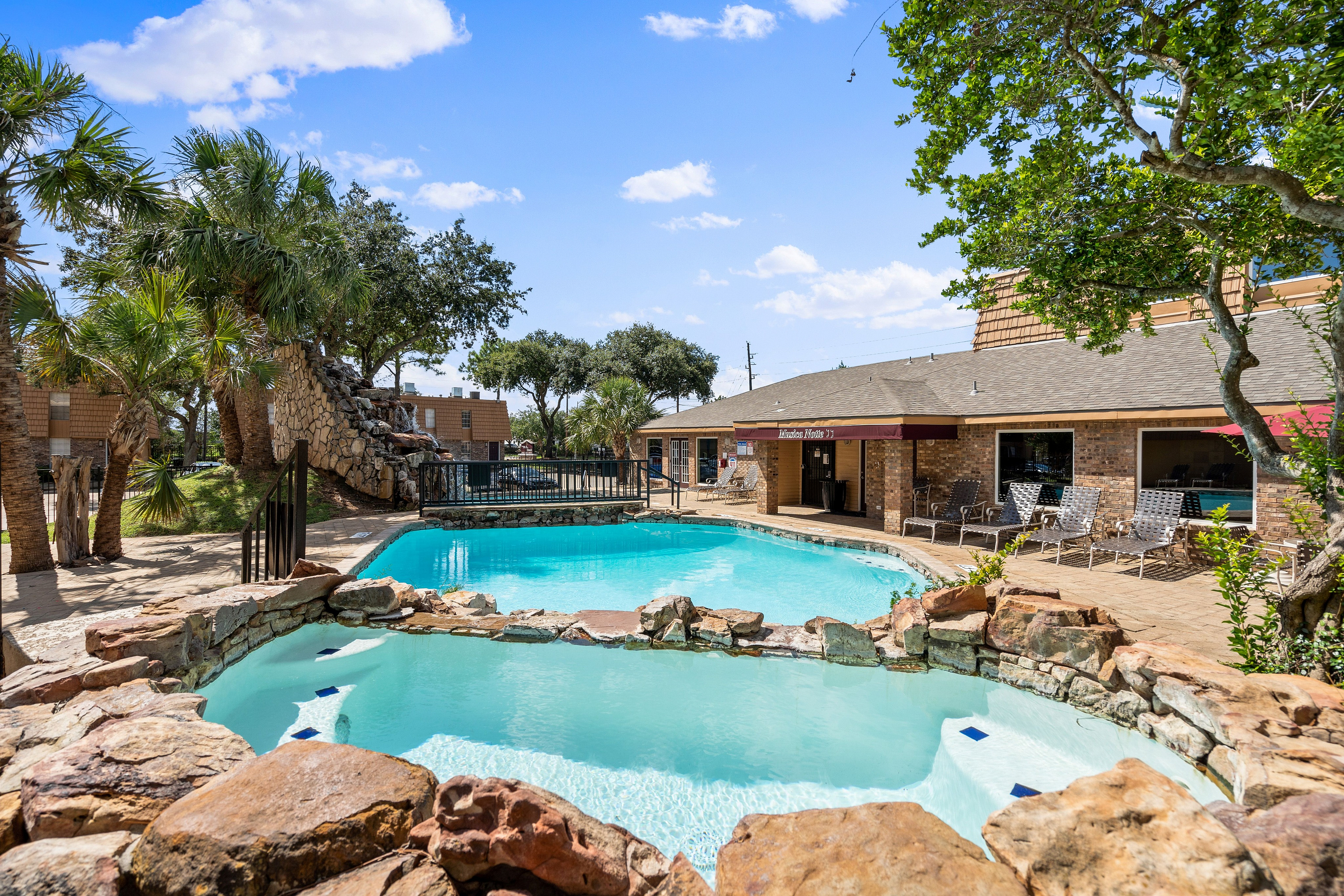 Inviting apartment complex pool area with cascading waterfall, surrounded by lush greenery and modern lounge chairs.
