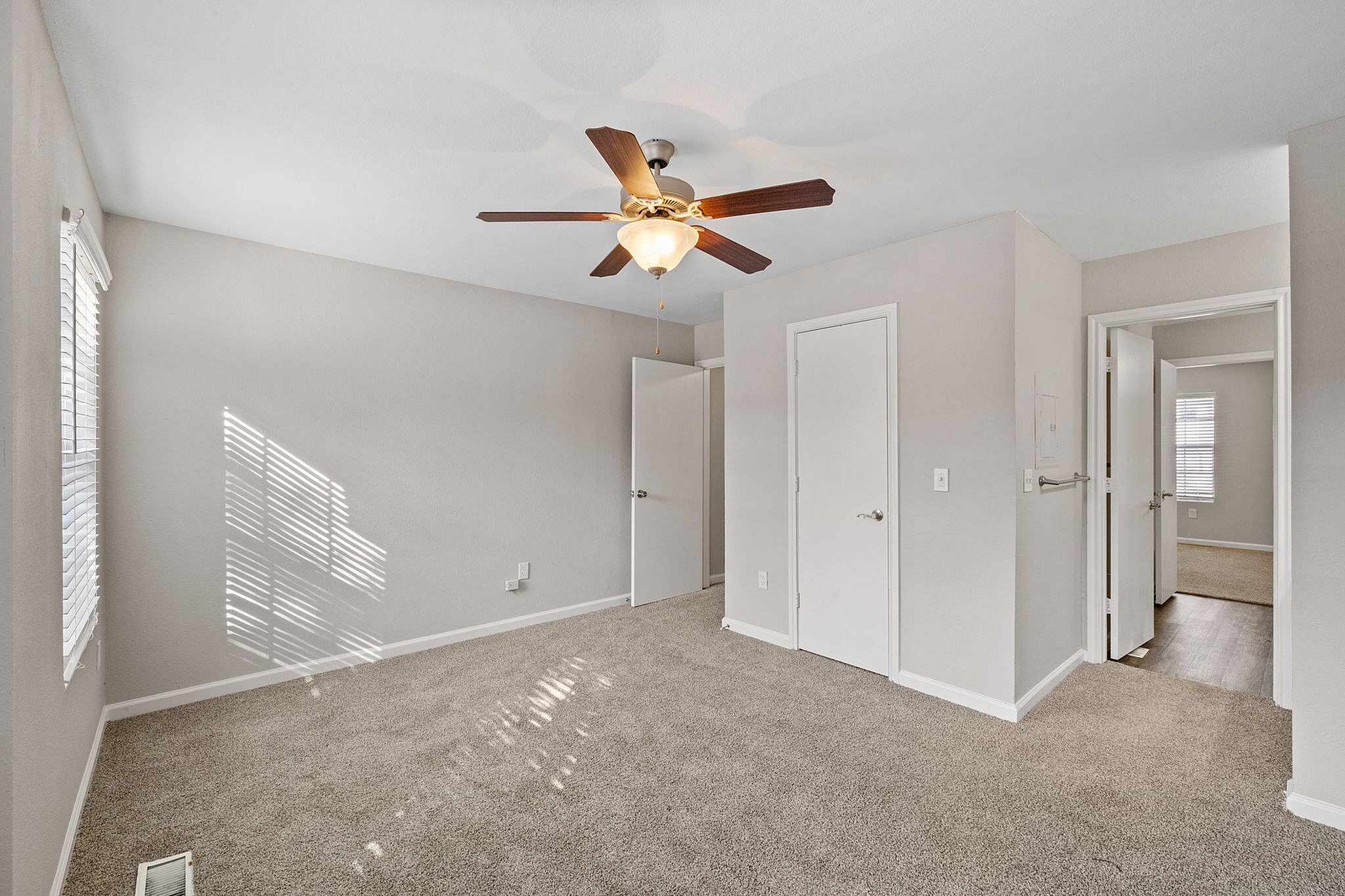 Bright and airy bedroom featuring plush carpeting, neutral walls, and a modern ceiling fan. Natural light streams through large windows creating a warm, inviting space.