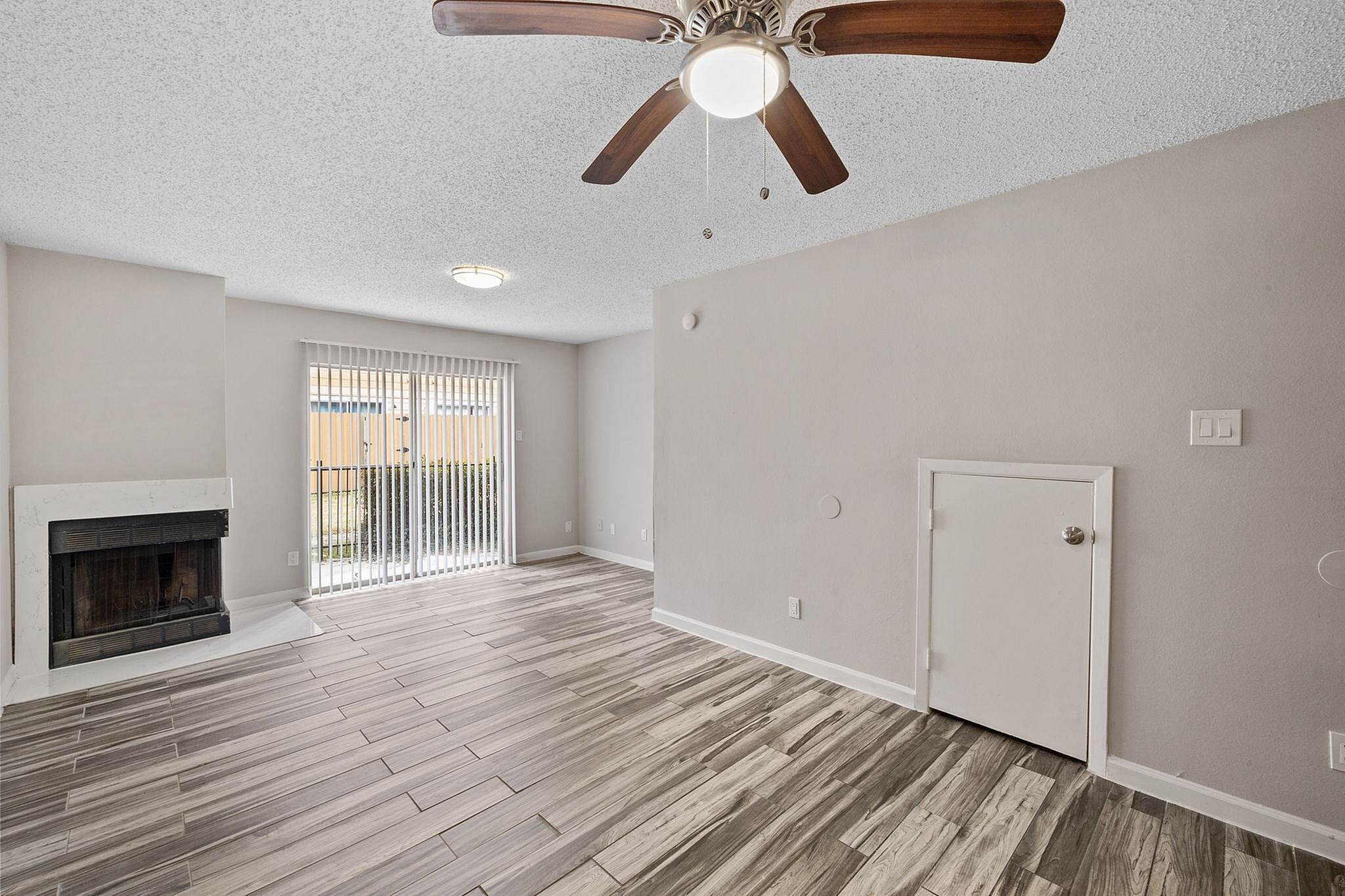 Inviting living room with modern wood flooring, ceiling fan, and cozy fireplace, leading to a bright patio through elegant sliding glass doors.