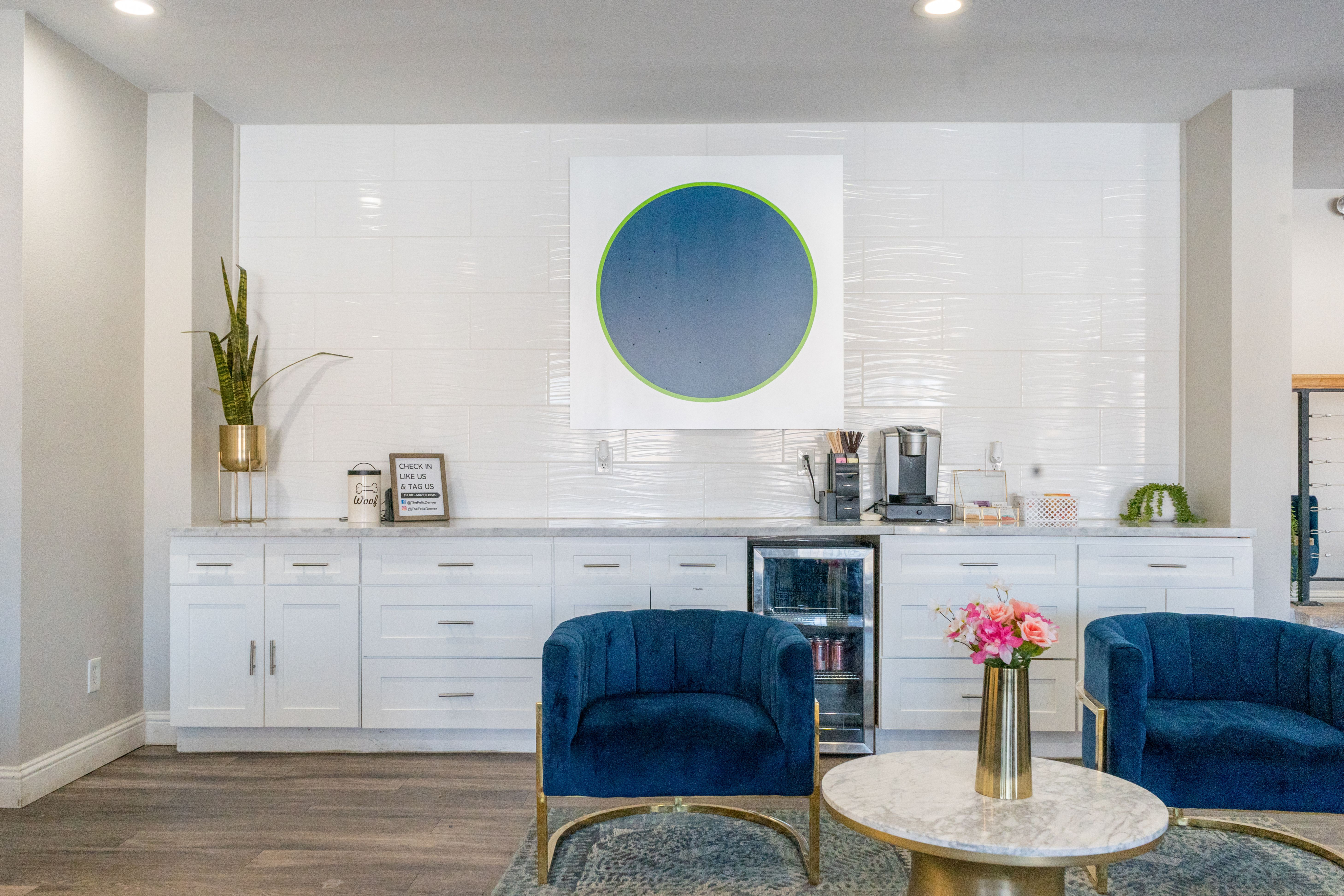 Elegant modern lounge area with plush blue chairs, chic marble table, and stylish kitchenette featuring sleek cabinetry and artful decor.