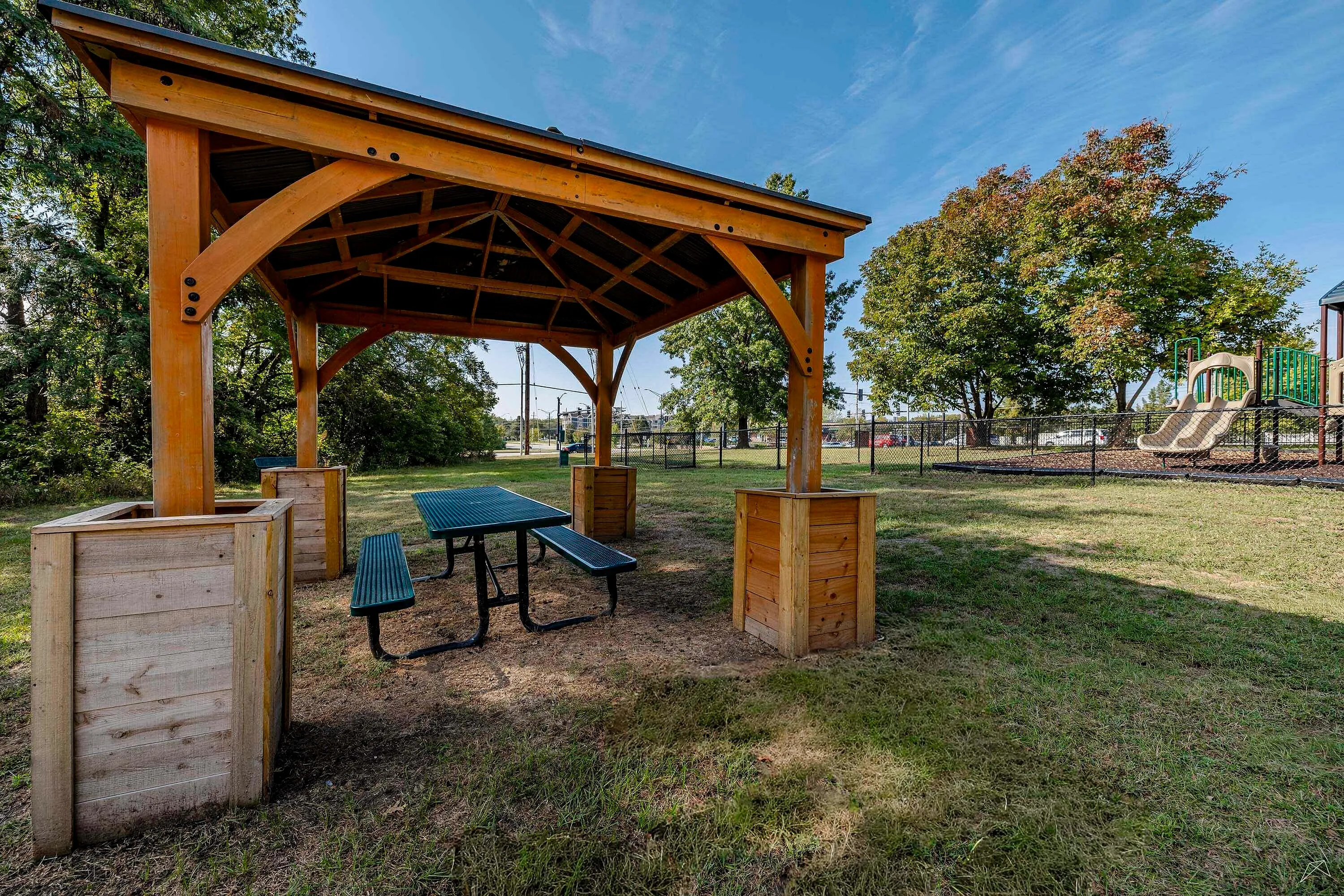 Inviting outdoor gazebo with picnic table in a lush park setting, adjacent to a vibrant playground under a clear blue sky.