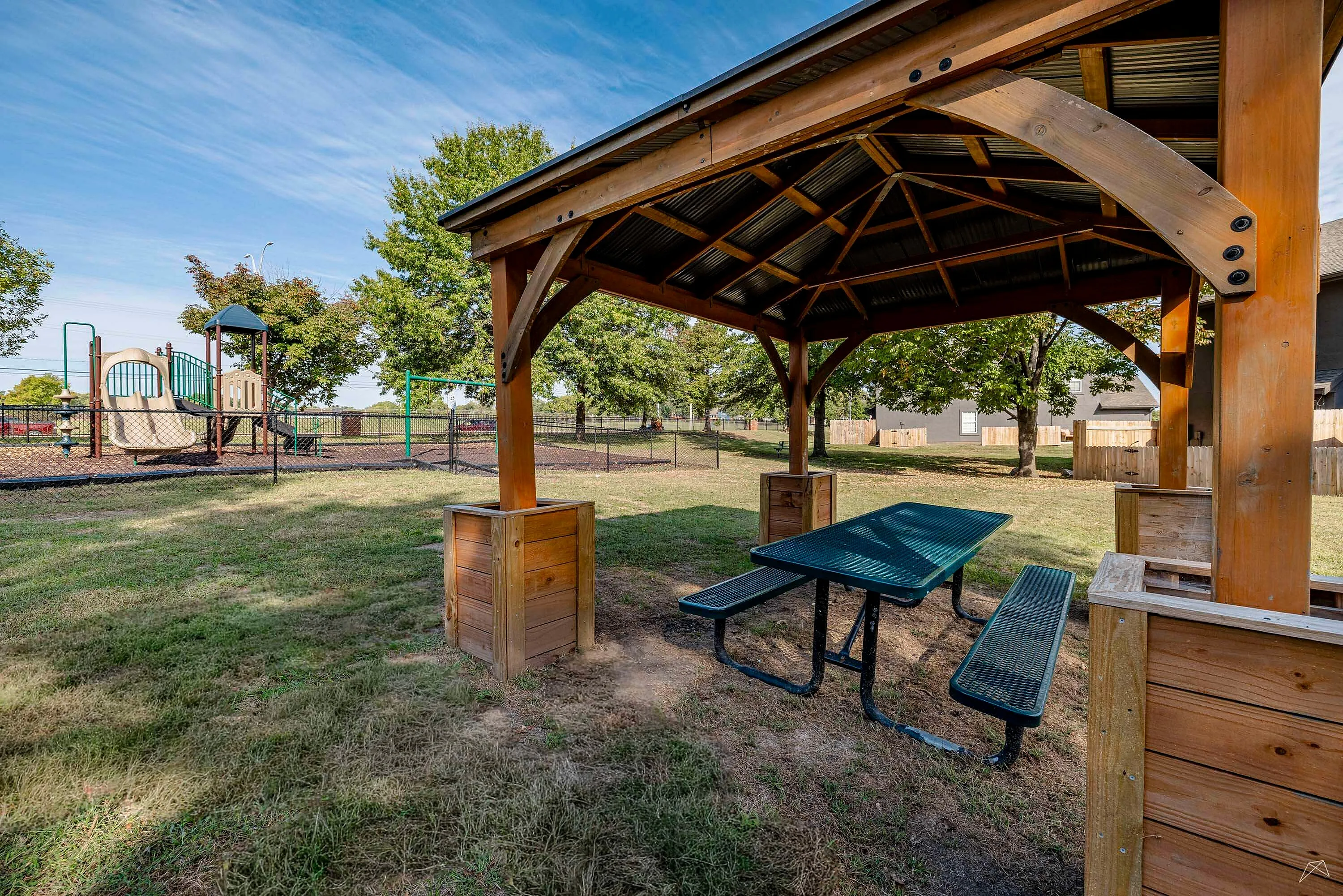 Charming outdoor space showcasing a wooden pavilion with picnic tables, perfect for family gatherings, next to a fenced playground surrounded by lush greenery.