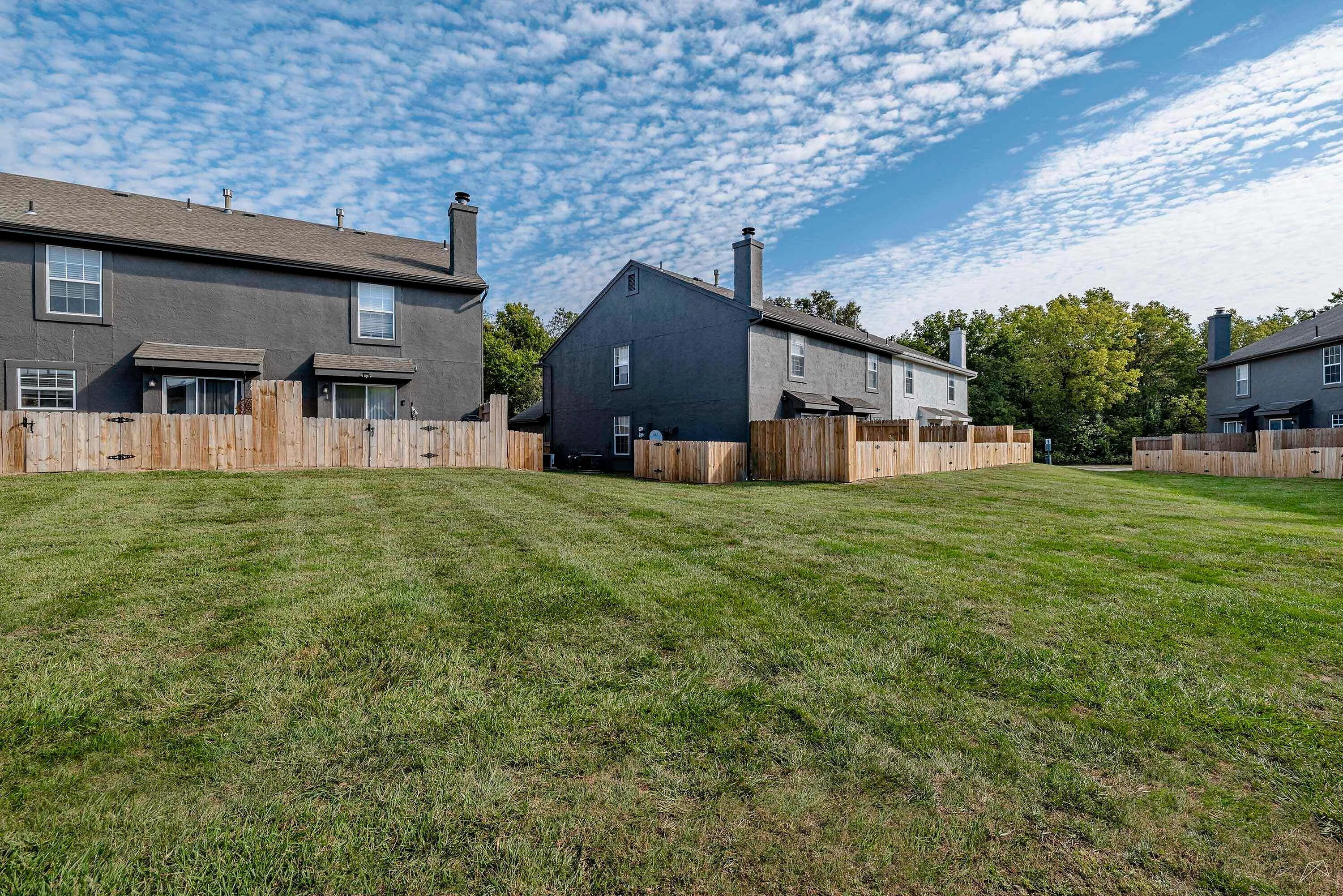 Spacious backyard view of modern townhomes featuring fenced patios, lush green lawn, and a clear blue sky.
