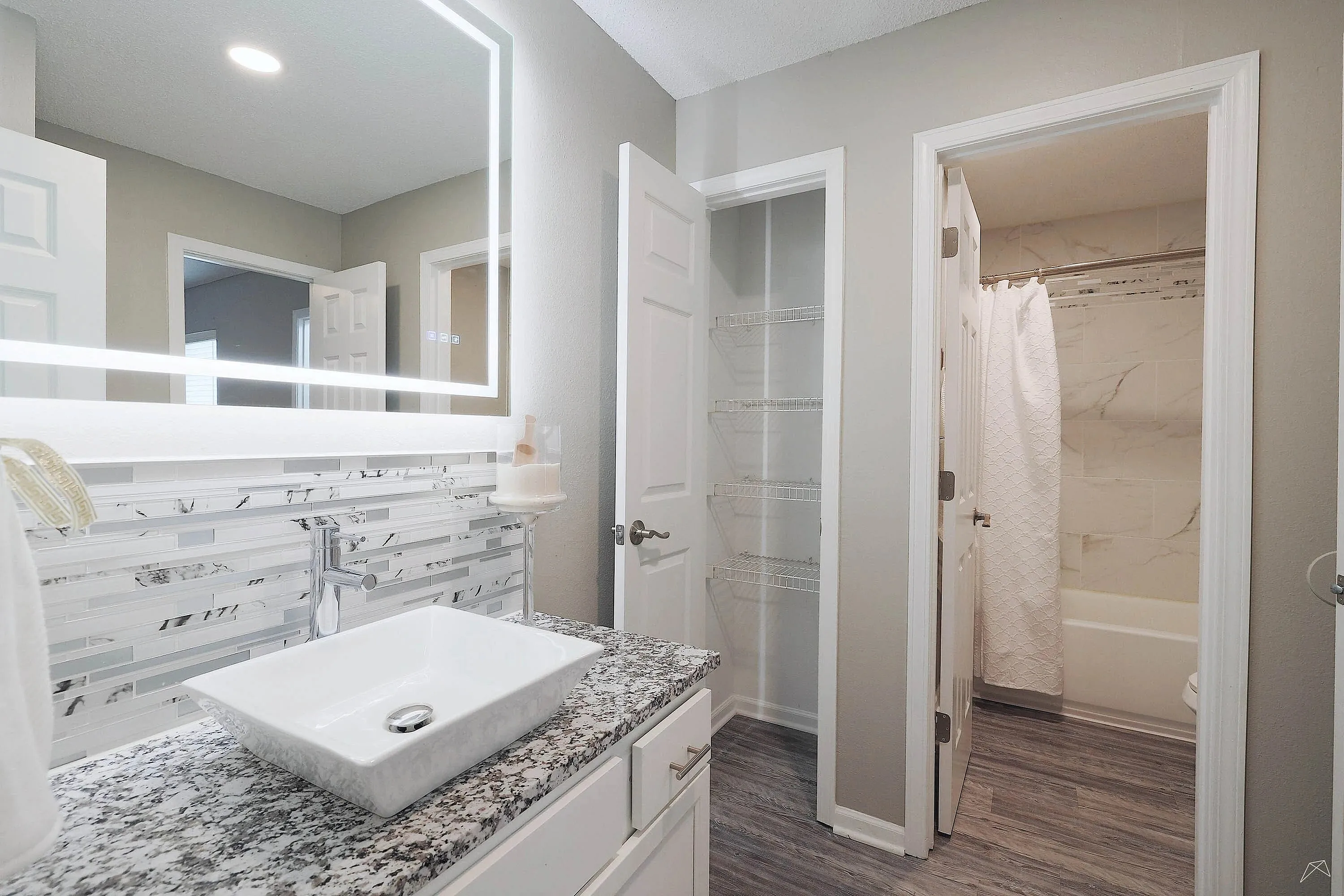 Elegant bathroom with modern granite countertop, illuminated mirror, and luxurious shower. Spacious design with sleek storage shelves.