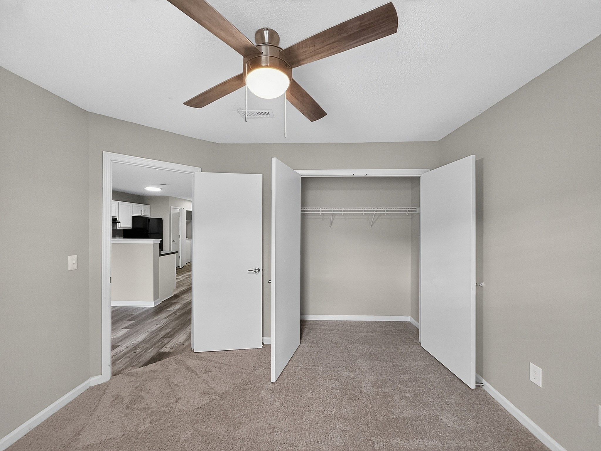Inviting bedroom featuring a modern ceiling fan, plush carpeting, and access to a bright hallway leading to the kitchen with stylish flooring.