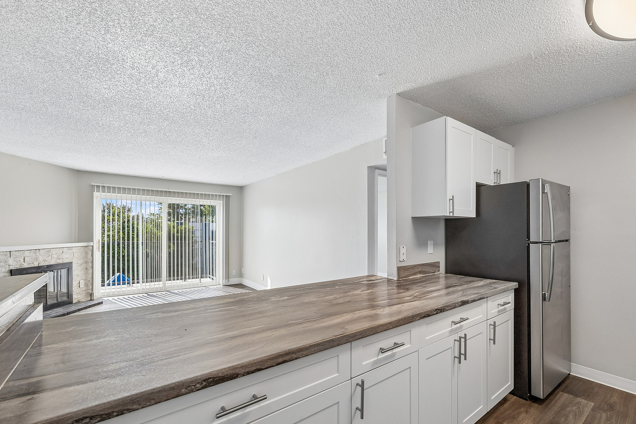 Bright living area with modern kitchen, featuring sleek countertops, stainless steel fridge, and large sliding glass doors opening to balcony with lush green views.