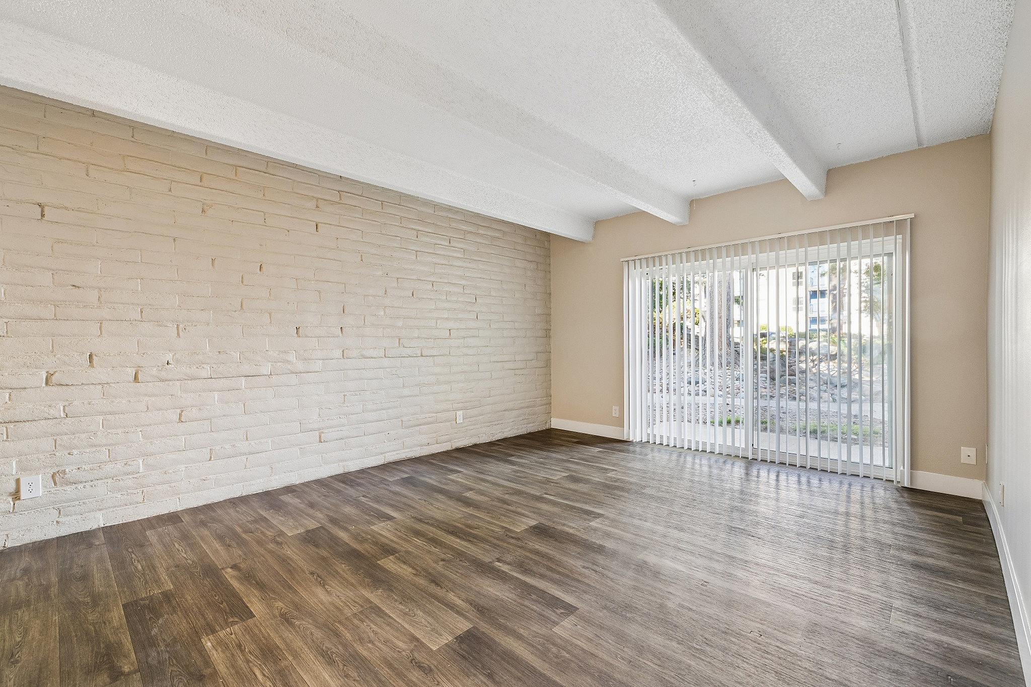 Bright and airy living room with wood-style flooring, exposed brick accent wall, and floor-to-ceiling sliding glass doors leading to a scenic outdoor view.