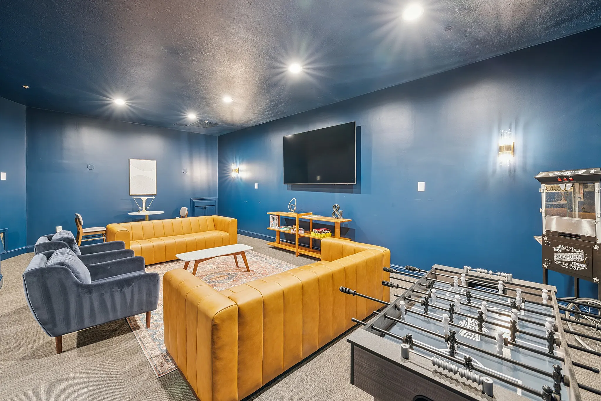 Chic entertainment room with modern blue and mustard sofas, large TV, foosball table, and popcorn machine, ideal for leisure and social gatherings.