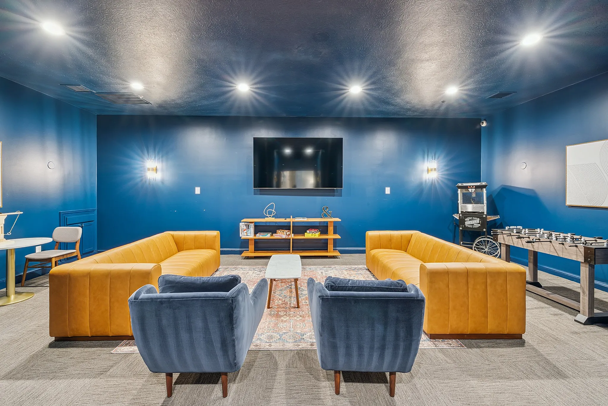 Chic entertainment room with vibrant blue walls, plush mustard sofas, a wall-mounted TV, and a sleek foosball table for ultimate relaxation and fun.