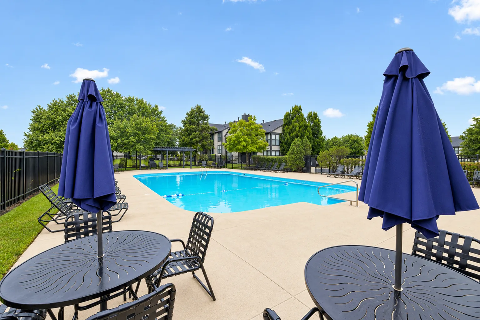 Inviting outdoor pool area with lounging chairs and blue umbrellas, surrounded by lush greenery and modern apartments.