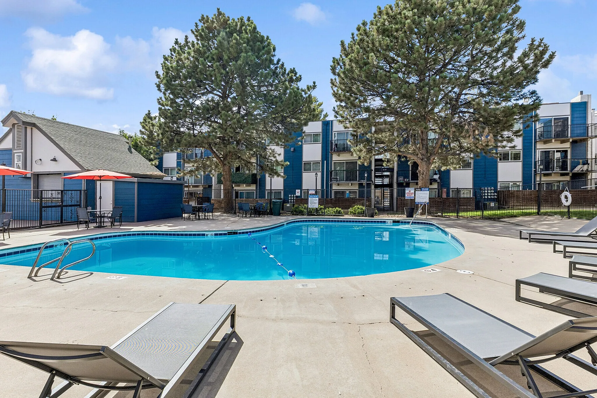 Inviting apartment complex with a sparkling outdoor pool, surrounded by lounge chairs, tall trees, and modern buildings with balconies.