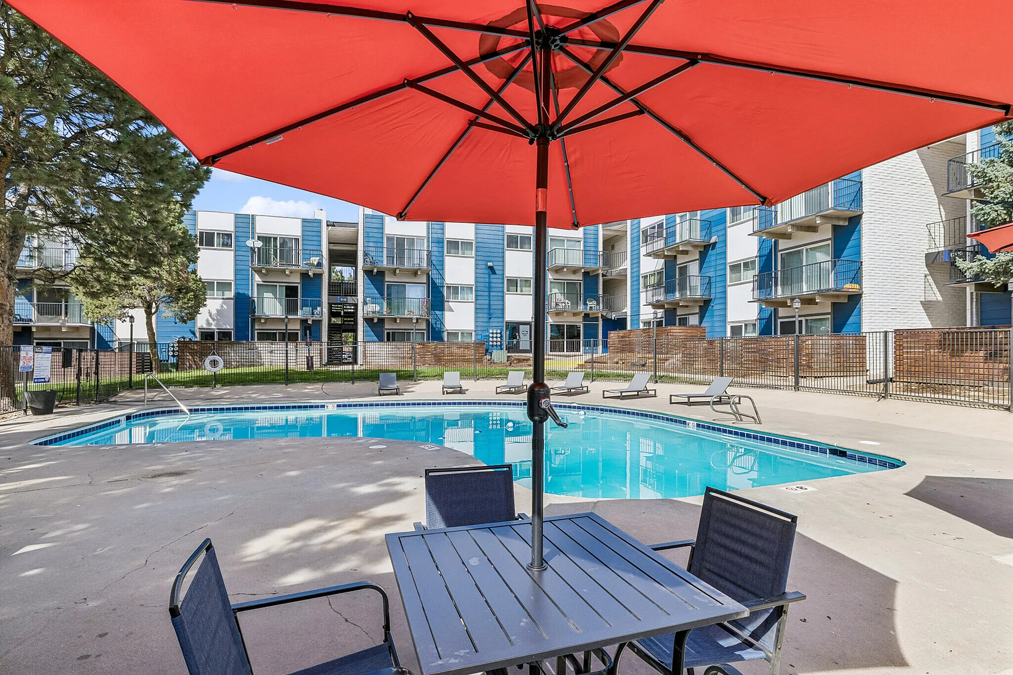 Inviting apartment complex with a sparkling pool, stylish lounge chairs, and vibrant red umbrellas in a modern courtyard setting.