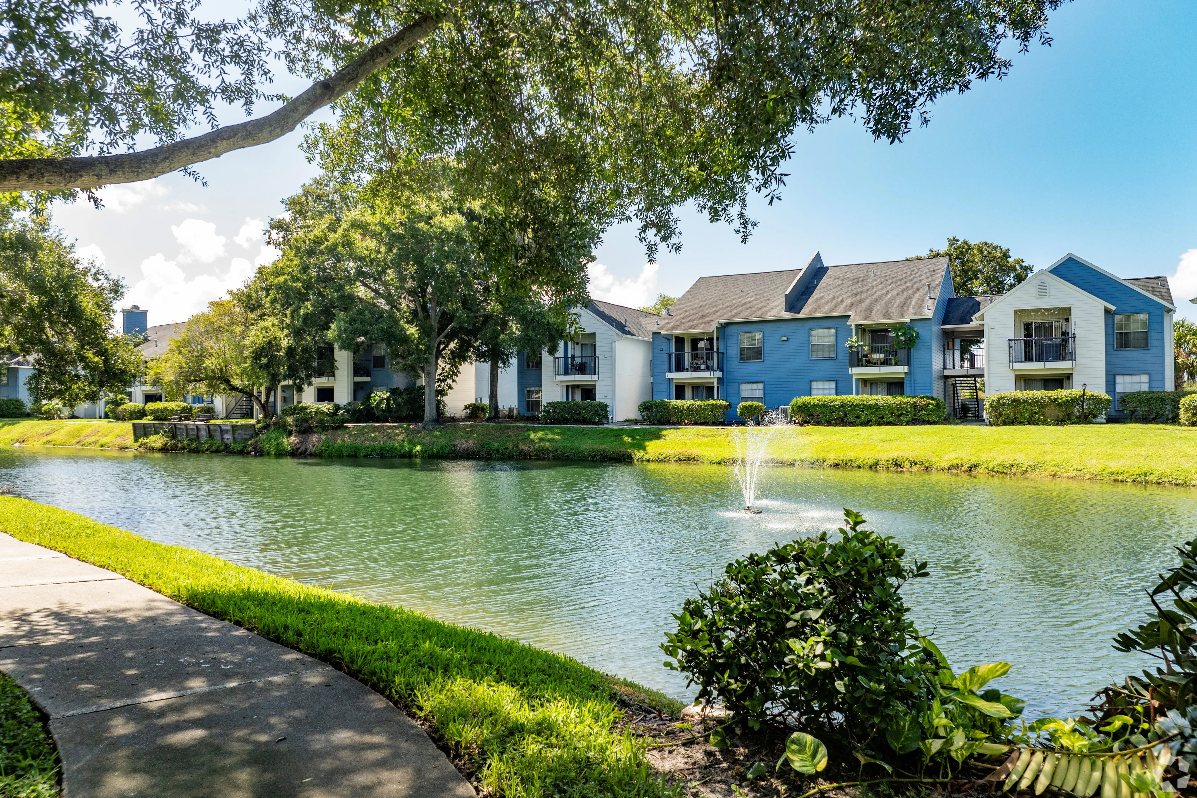 Charming lakeside apartment complex with blue and white buildings, surrounded by lush greenery and a serene pond featuring a decorative fountain.