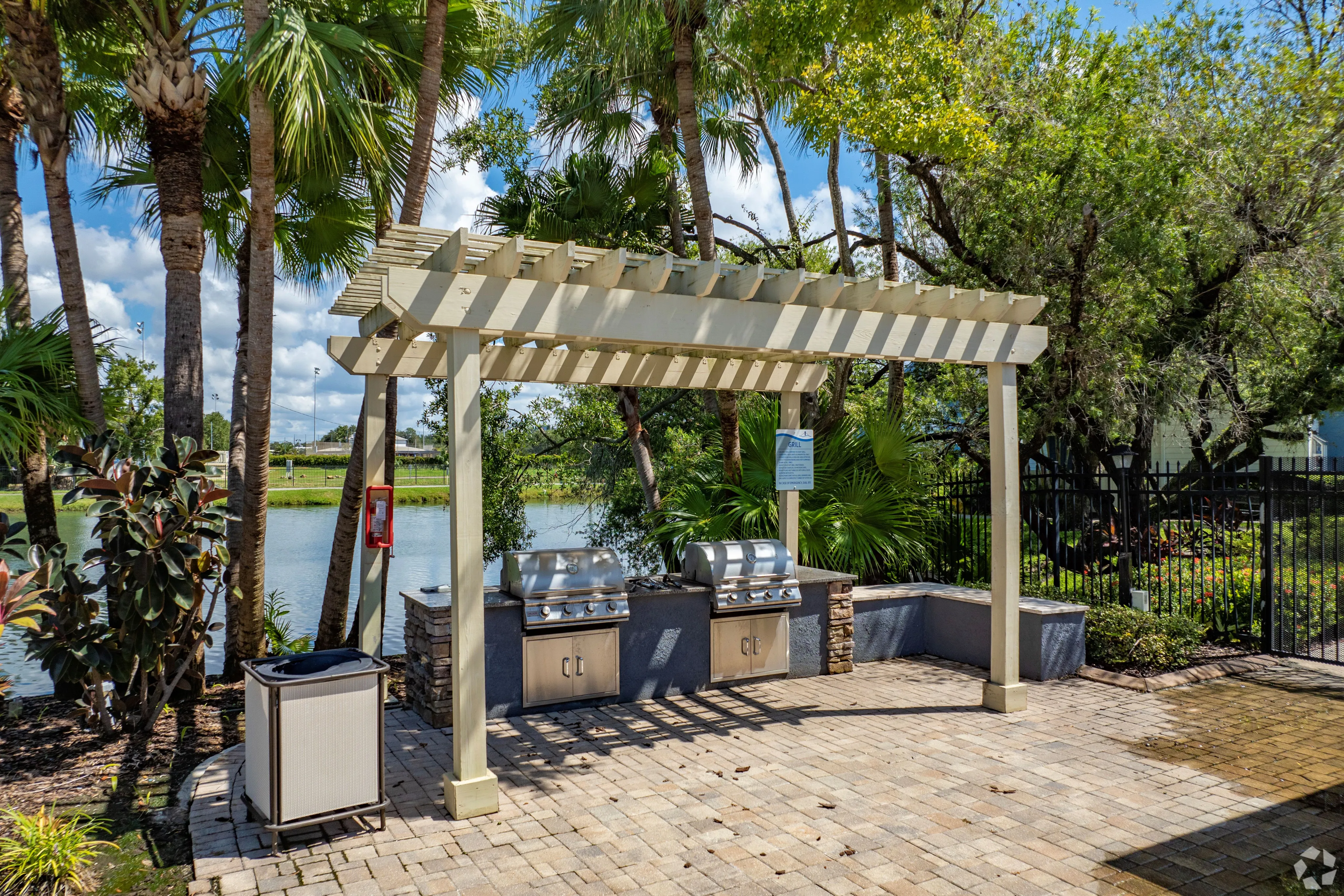 Charming outdoor grilling area with modern BBQ stations under a stylish pergola, surrounded by lush palm trees and a tranquil water view.