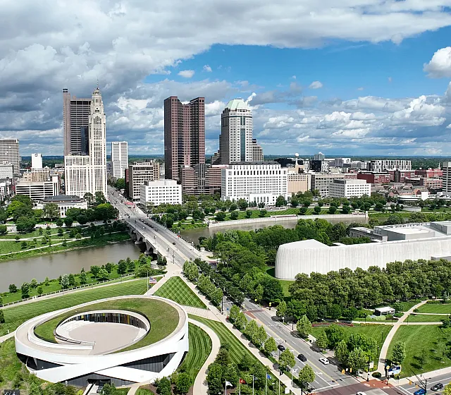 Vibrant cityscape with modern skyscrapers, lush green parkland, and a winding river under a bright sky.