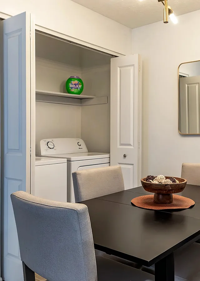 Chic apartment dining area with a sleek black table, surrounded by modern chairs and adjacent to a compact laundry nook with washer and dryer.
