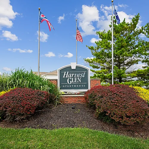 Welcoming entrance of Harvest Glen Apartments with vibrant landscaping and flags against a clear blue sky.