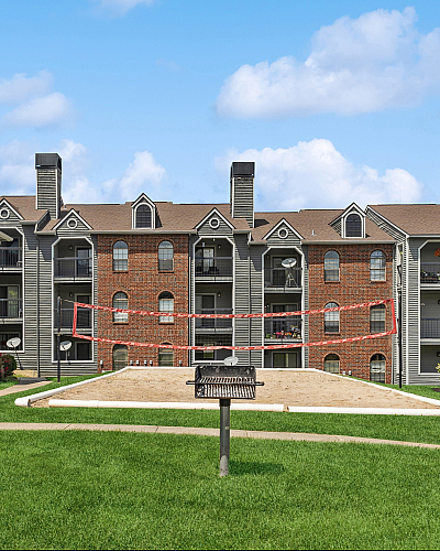Beautiful apartment complex featuring brick and siding, surrounded by lush greenery and a volleyball court.