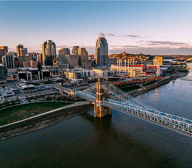 Aerial view of a city skyline with a prominent suspension bridge crossing a river at sunset.
