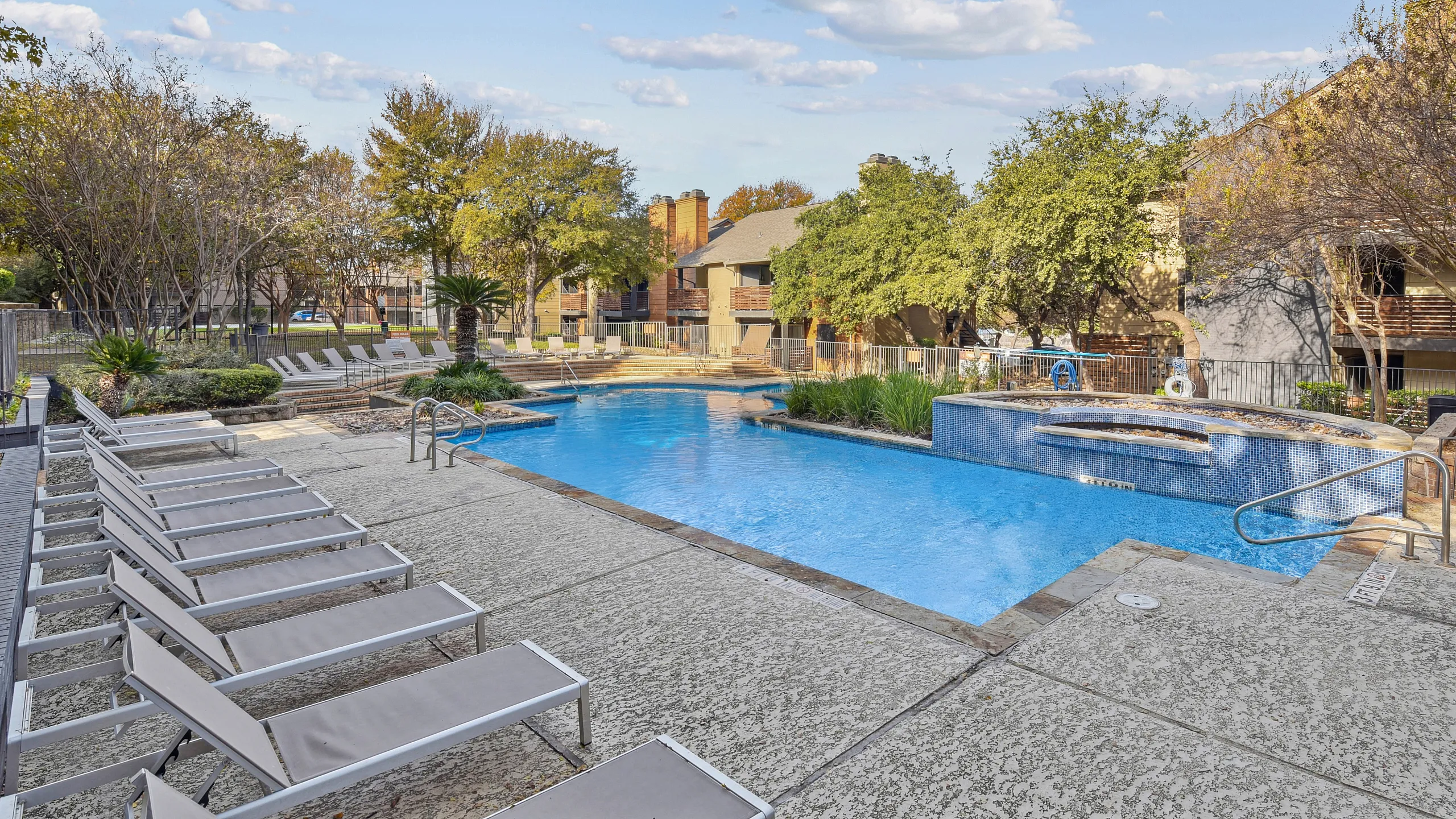 Inviting apartment complex pool area with luxurious lounge chairs, surrounded by lush greenery and vibrant blue skies.