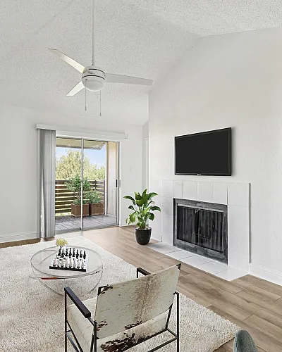 Bright and modern living room with elegant decor, large sliding glass doors leading to a balcony with lush greenery, a cozy fireplace, and stylish furnishings including a leather sofa and glass coffee table.