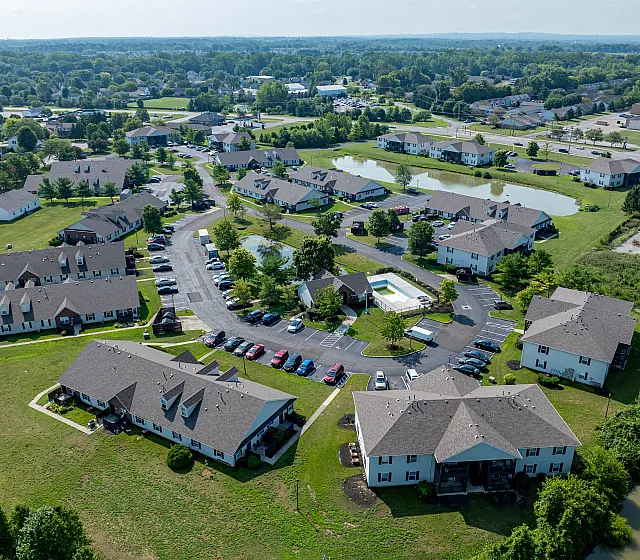 Aerial view of charming community layout featuring spacious apartment buildings surrounded by lush greenery, a serene pond, and a refreshing swimming pool.