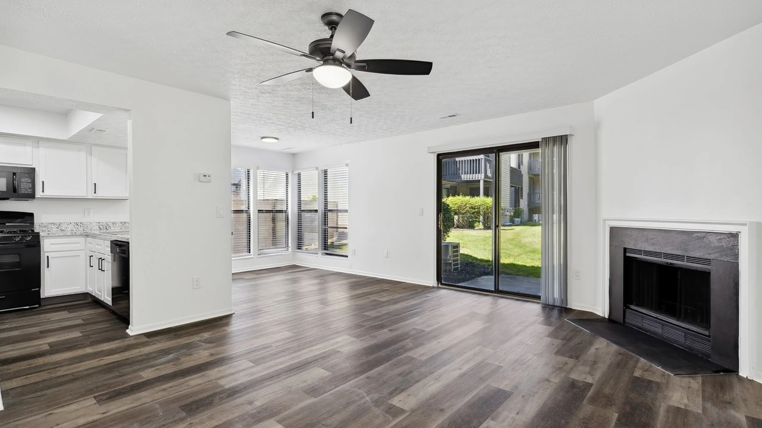 Bright and modern open-concept living area with wood flooring, ceiling fan, and sliding glass doors leading to a lush outdoor space. Features a stylish kitchen with sleek black appliances.