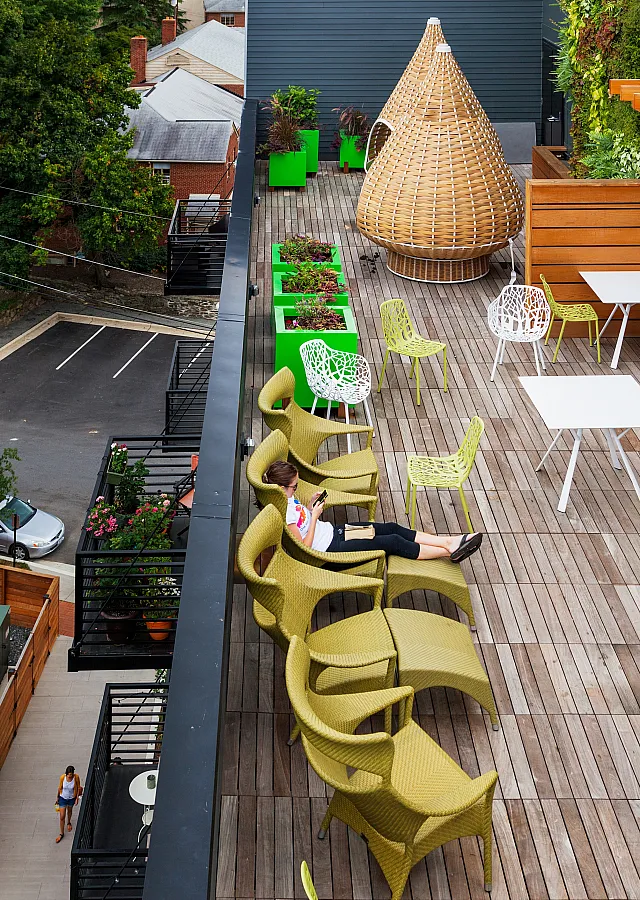 Chic urban rooftop patio with wooden decking, vibrant green planters, stylish seating, and a cozy wicker pod, offering a relaxing outdoor escape.