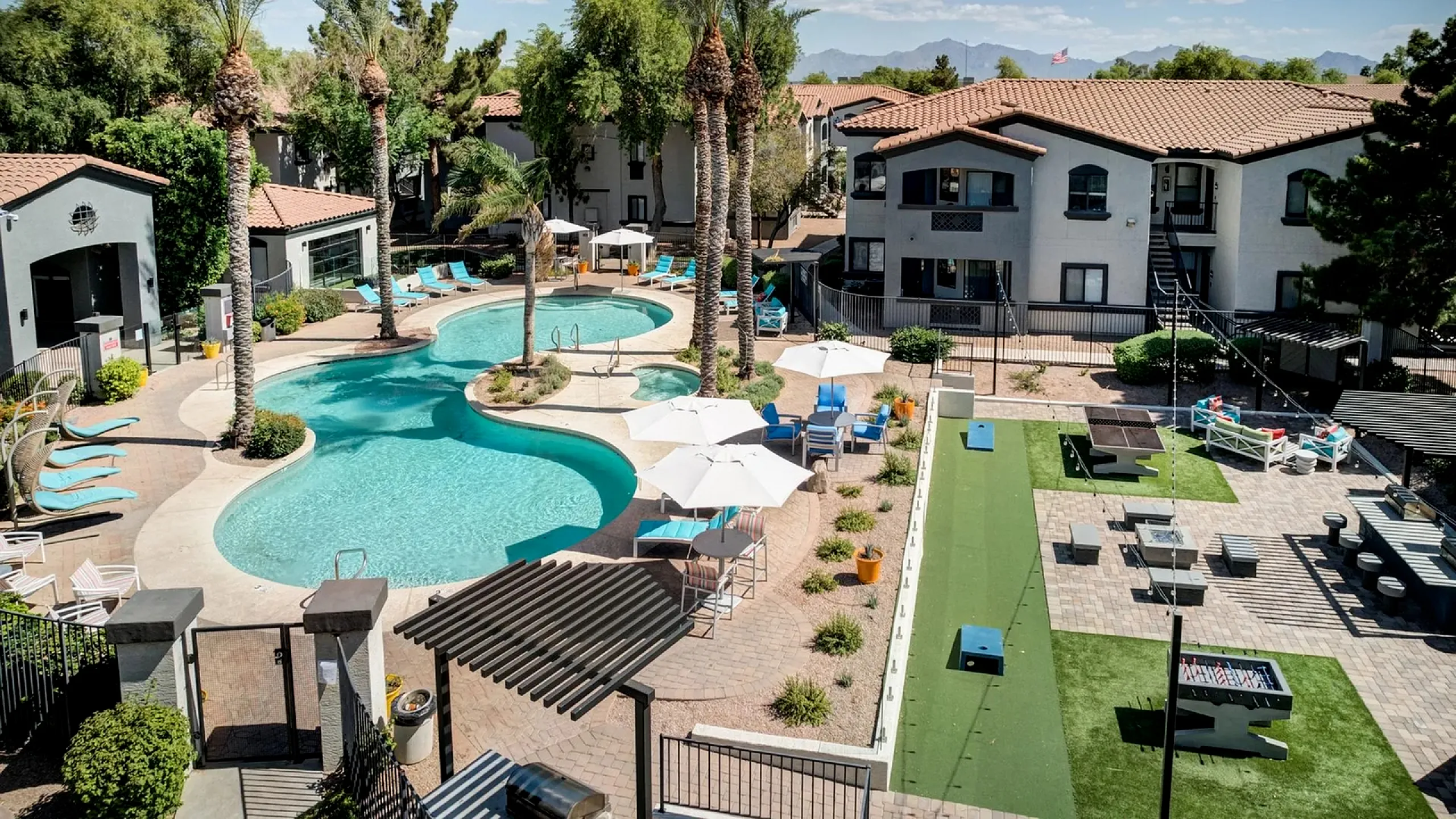 Luxurious apartment complex featuring a stunning pool with palm trees, relaxing lounge chairs, and vibrant outdoor seating areas, set against a backdrop of picturesque mountains.