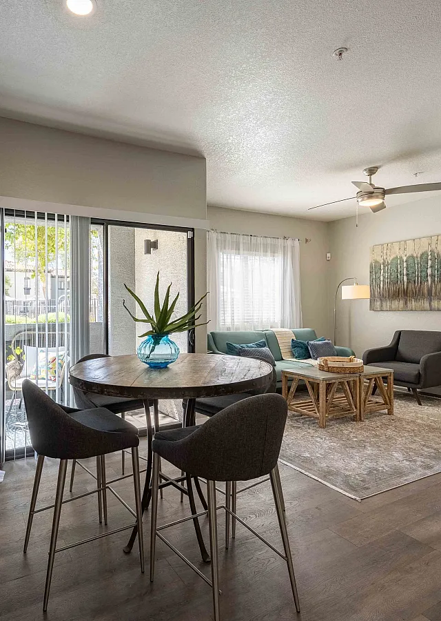 Elegant apartment living room with stylish decor, featuring a cozy seating area, a round wooden dining table, and lush greenery. Natural light flows through large sliding glass doors, creating a bright and inviting atmosphere.