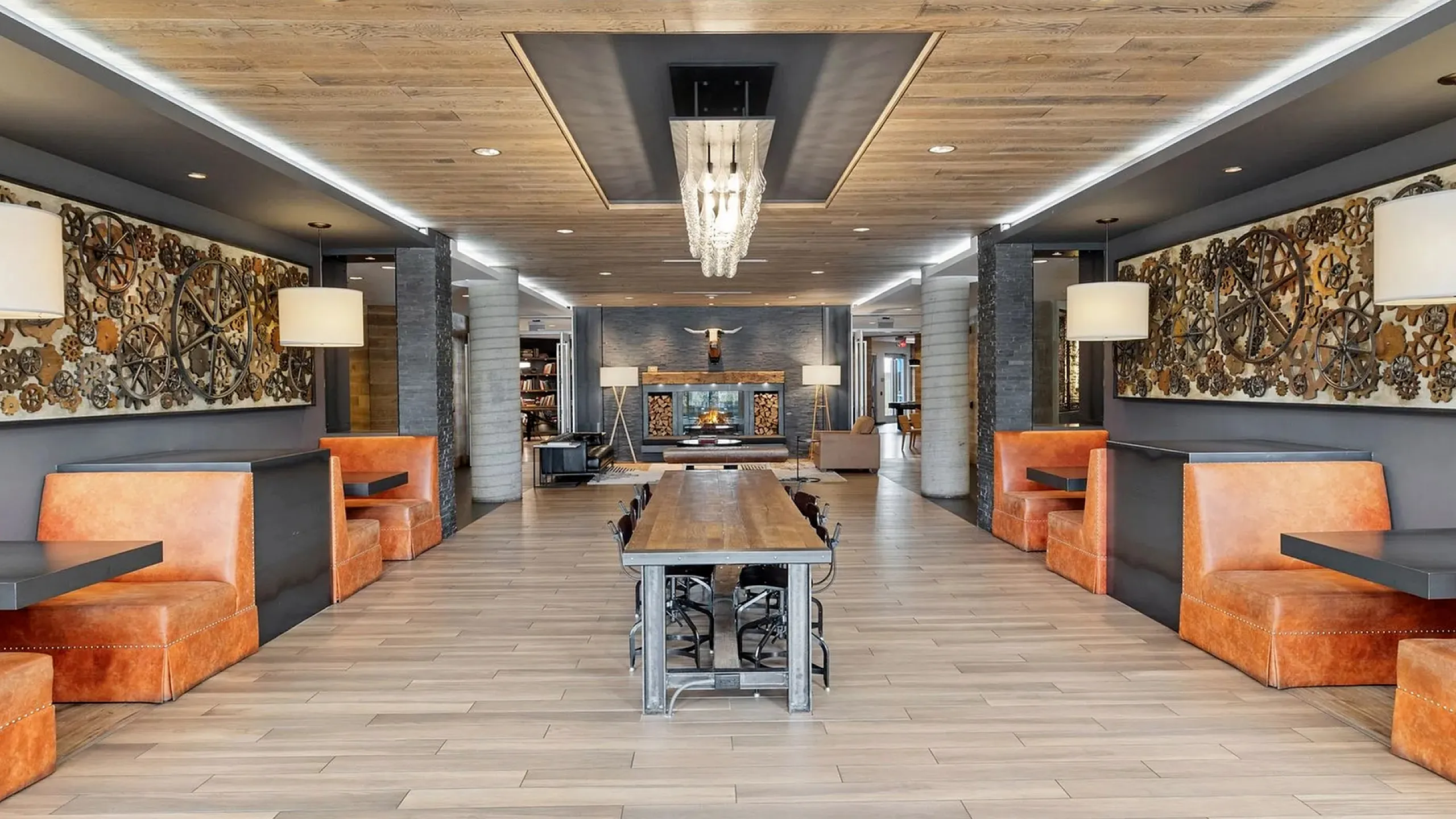 Modern lounge area with rustic wooden flooring, stylish orange booths, and a striking chandelier, perfect for social gatherings.
